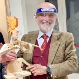 Uomo anziano che indossa una visiera protettiva e tiene in mano una scultura in legno di una figura con capelli di fiamma.