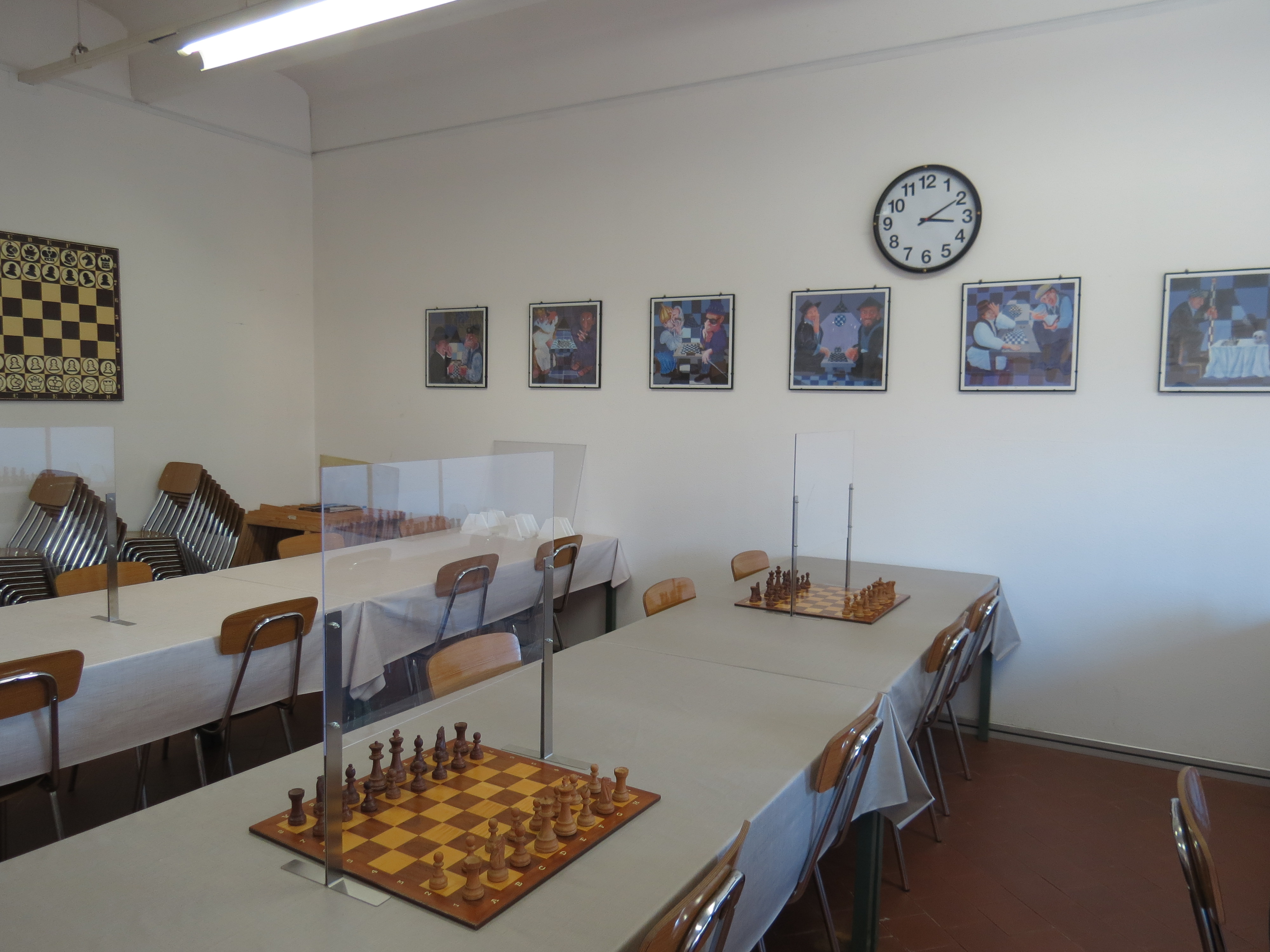 Interno di una sala contenente un tavolo da scacchi, sedie e una serie di foto appese al muro. Una sveglia è visibile sopra il tavolo.