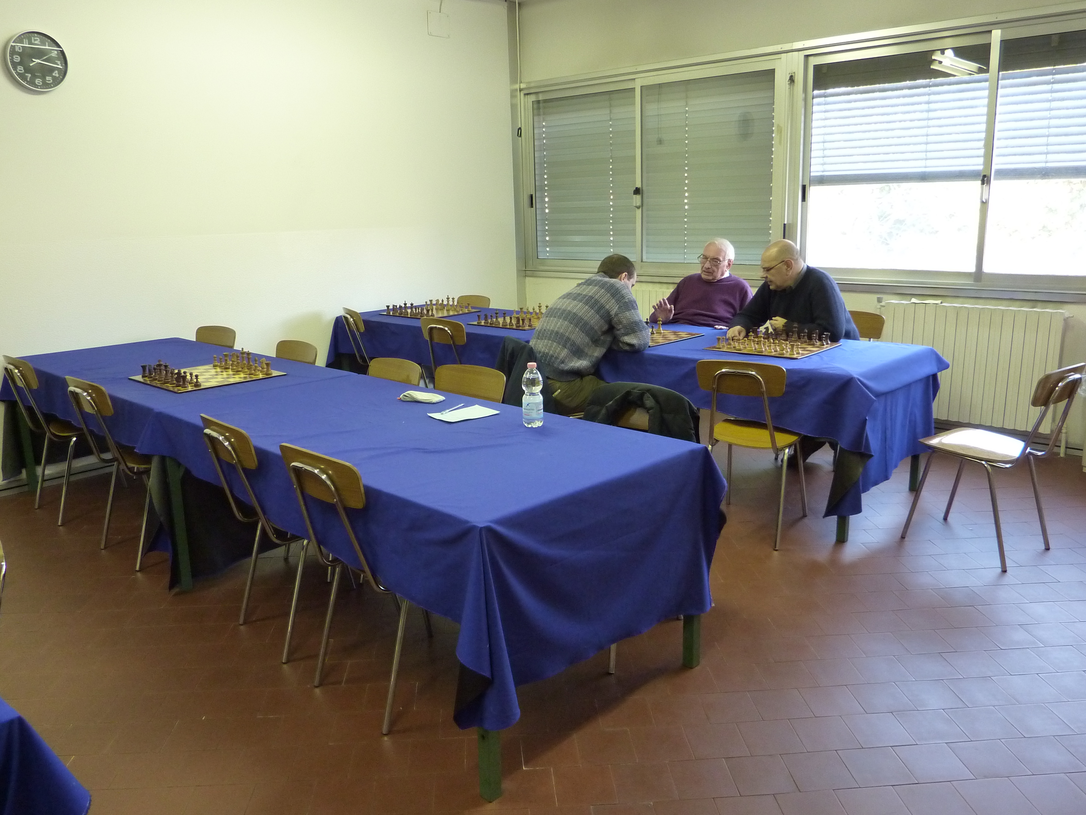 Interno della sala del Circolo di scacchi con tavoli coperti da tovaglie blu e giocatori che si sfidano a scacchi.