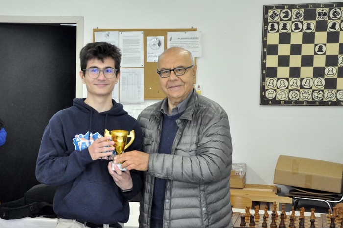 Un giovane giocatore di scacchi riceve un trofeo da un uomo anziano all'interno di un circolo scacchistico, con un'illustrazione di scacchi sul muro dietro di loro.