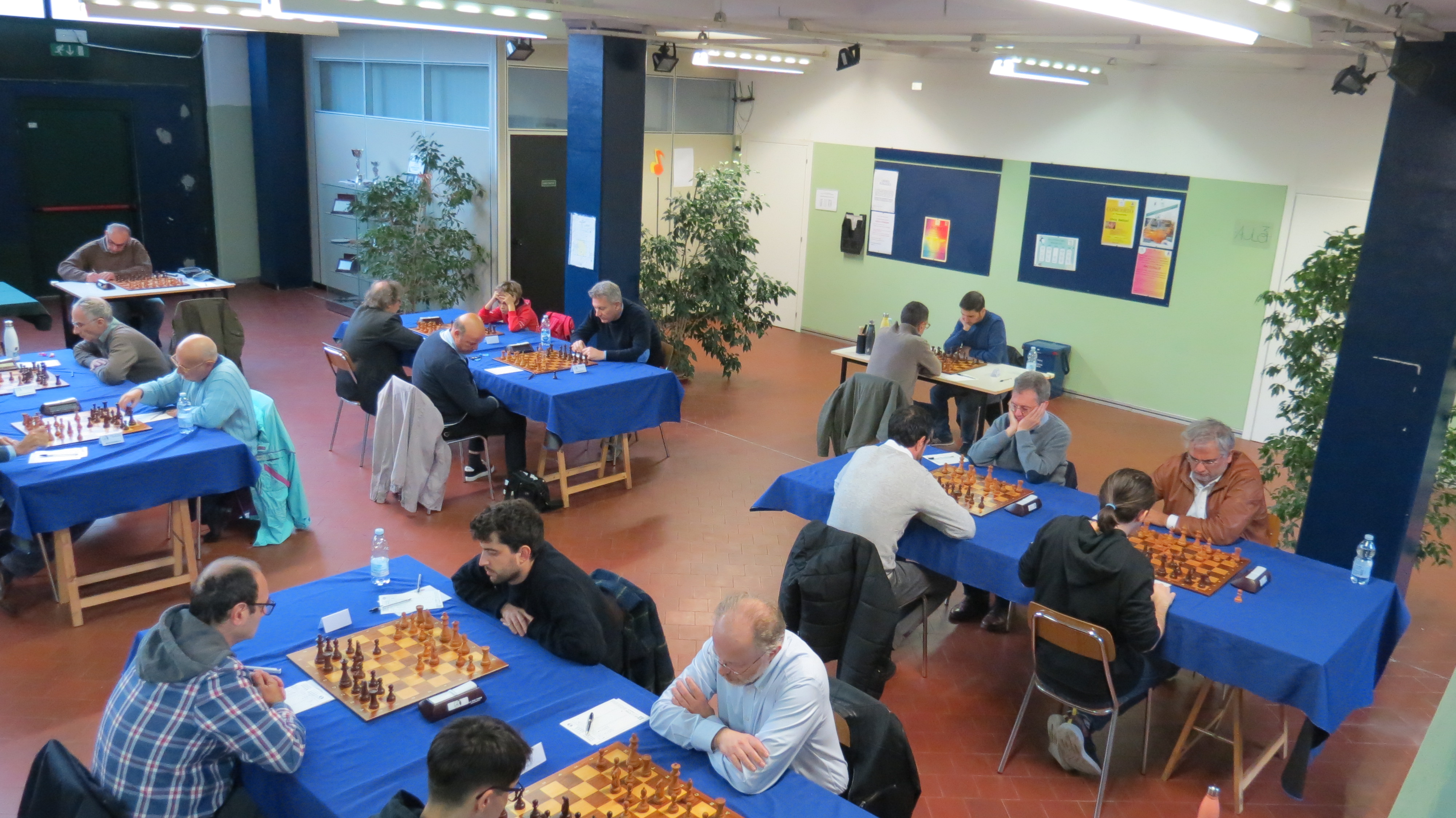 Vista aerea del torneo di scacchi con partecipanti seduti a tavoli blu mentre giocano e annotano le mosse.