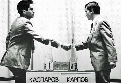 Garry Kasparov e Anatoly Karpov si stringono la mano prima di un match di scacchi, con tavoli e bandiere sullo sfondo, simbolo della loro storica rivalità.