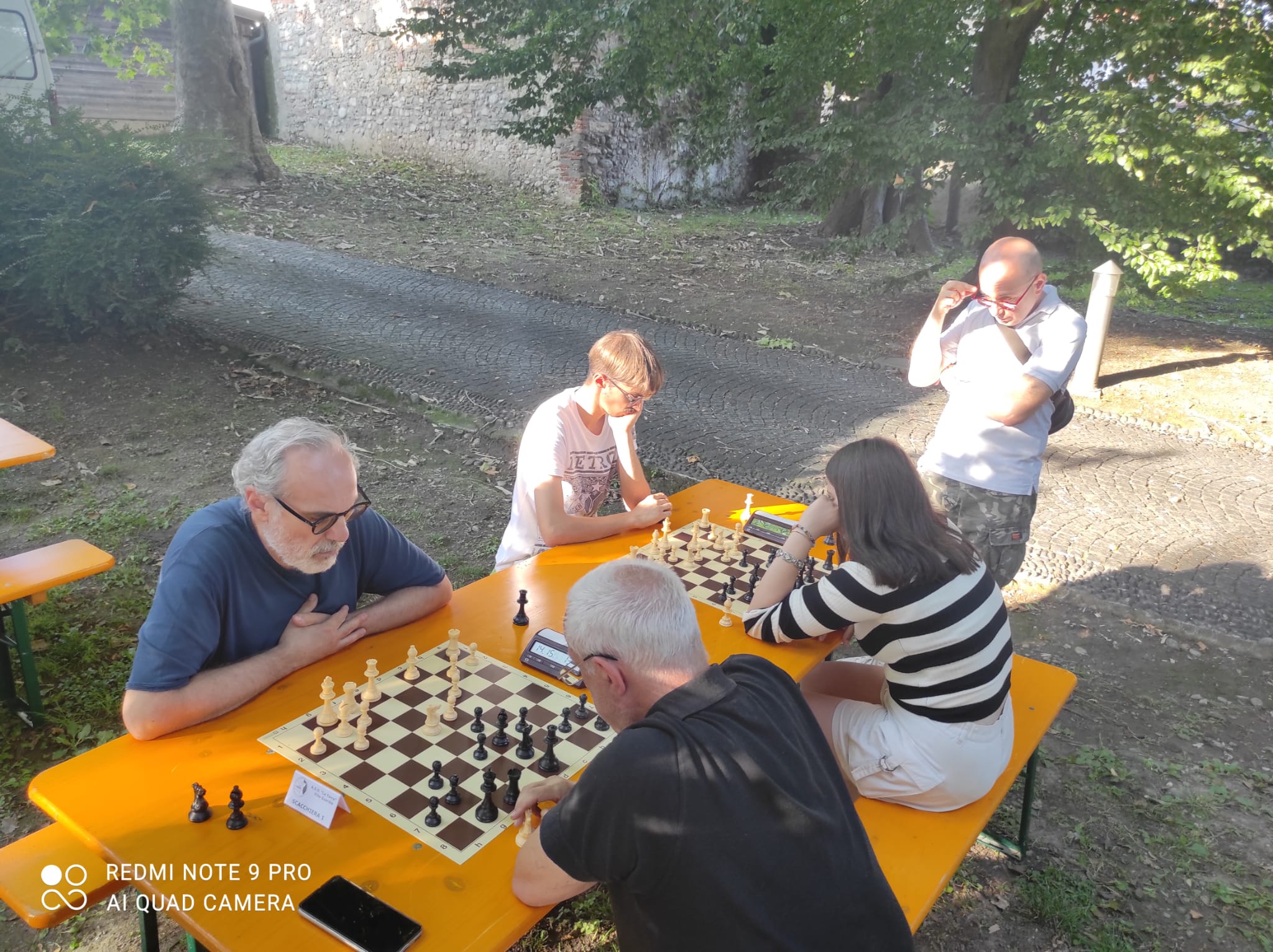 Giocatori impegnati in una partita di scacchi all'aperto, seduti attorno a un tavolo di legno giallo con una scacchiera.