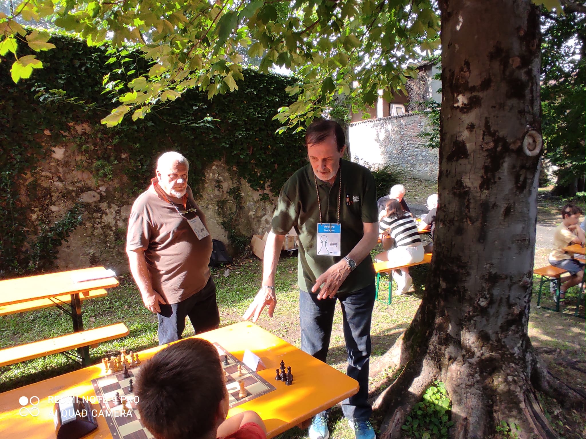 Due partecipanti discutono durante una partita di scacchi all'aperto, con un tavolo di gioco e sedie in legno visibili sullo sfondo.