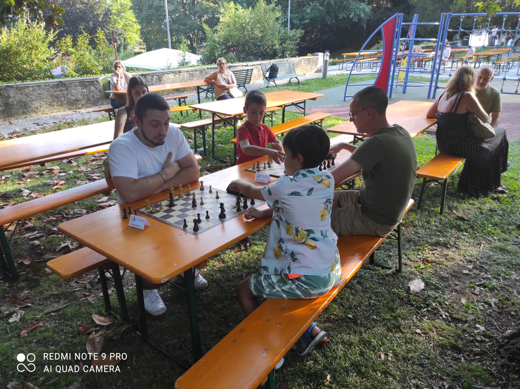 Giocatori di scacchi seduti attorno a tavoli nel parco durante un torneo, con un enfasi su bambini e adulti che partecipano alla competizione.
