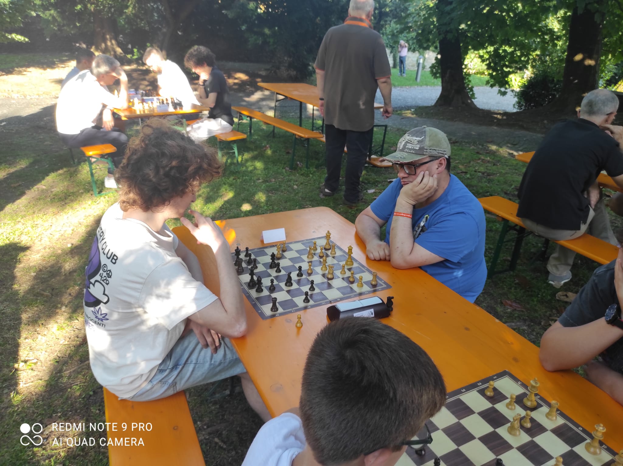 Due bambini giocano a scacchi su una tavola al parco, circondati da altri partecipanti del torneo.
