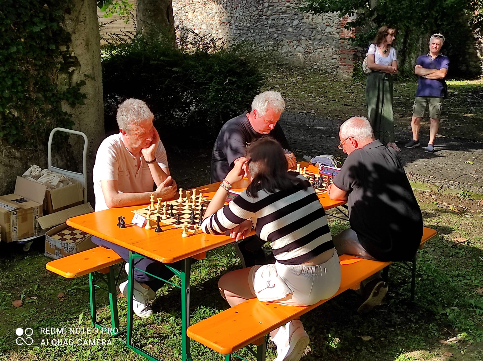Quattro giocatori di scacchi concentrati durante una partita all'aperto, seduti attorno a un tavolo in un'area verde.