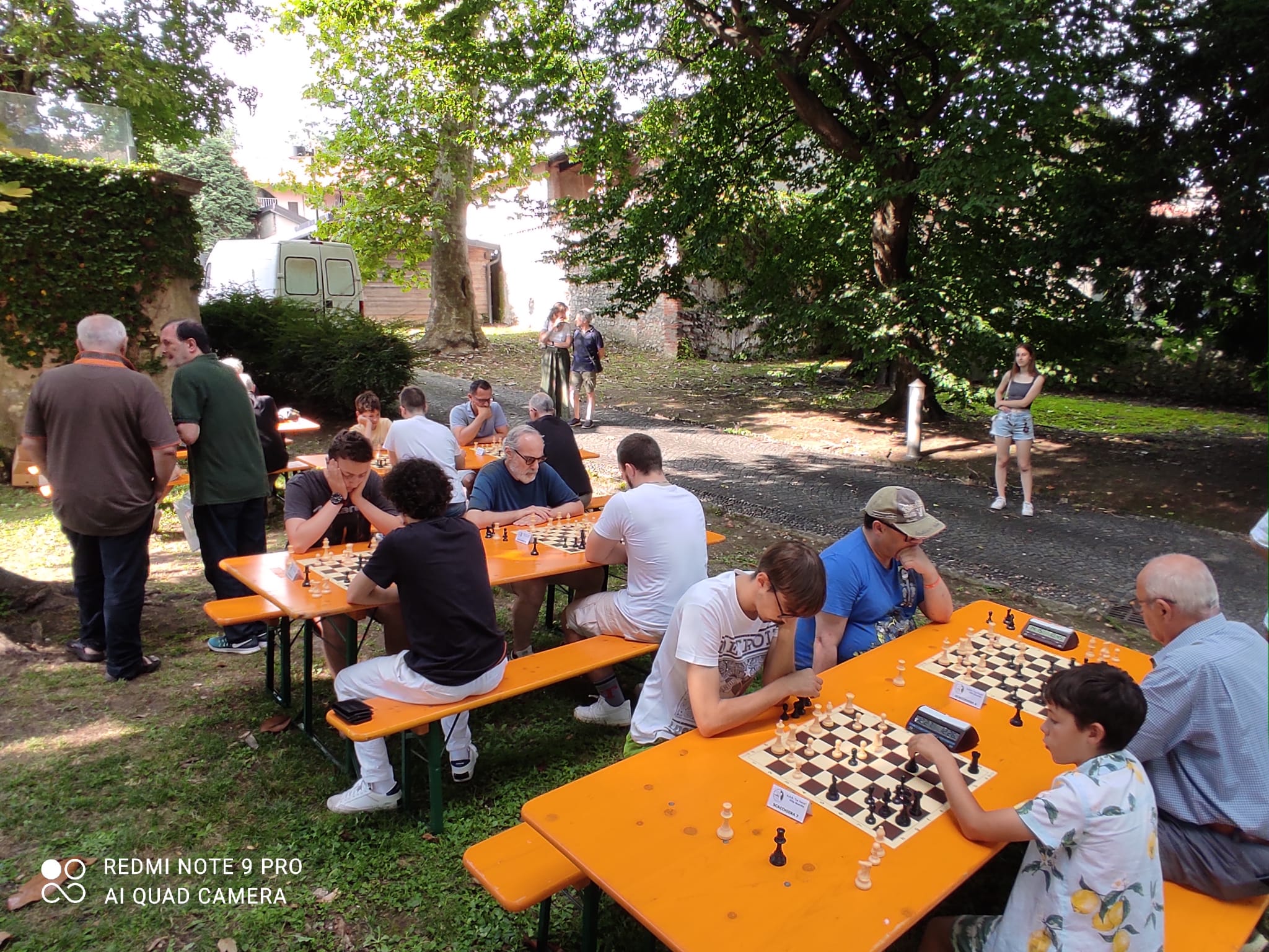 Un gruppo di giocatori di scacchi si sfida all'aperto, seduti a tavoli arancioni affollati nel Parco Comunale di Casnate con Bernate.