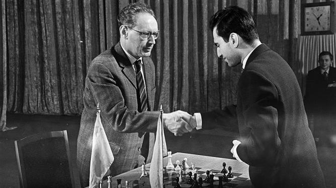 Due scacchisti si stringono la mano prima dell'inizio di una partita, con una scacchiera e pezzi di legno visibili davanti a loro.