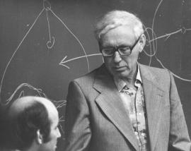 Due uomini conversano in una stanza con una lavagna sullo sfondo, sulla quale sono presenti disegni e diagrammi.