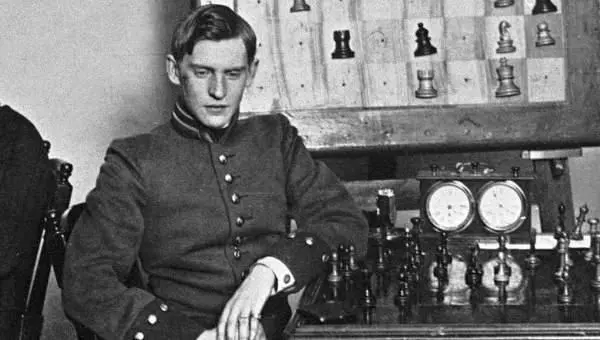 Un giovane Alexander Alekhine con un atteggiamento pensoso seduto accanto a una scacchiera, mentre i pezzi di scacchi e i cronometri sono visibili sul tavolo.