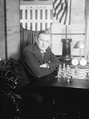 Un uomo seduto a una scrivania con una scacchiera e un orologio da scacchi, mentre le bandiere americane sono visibili sullo sfondo.