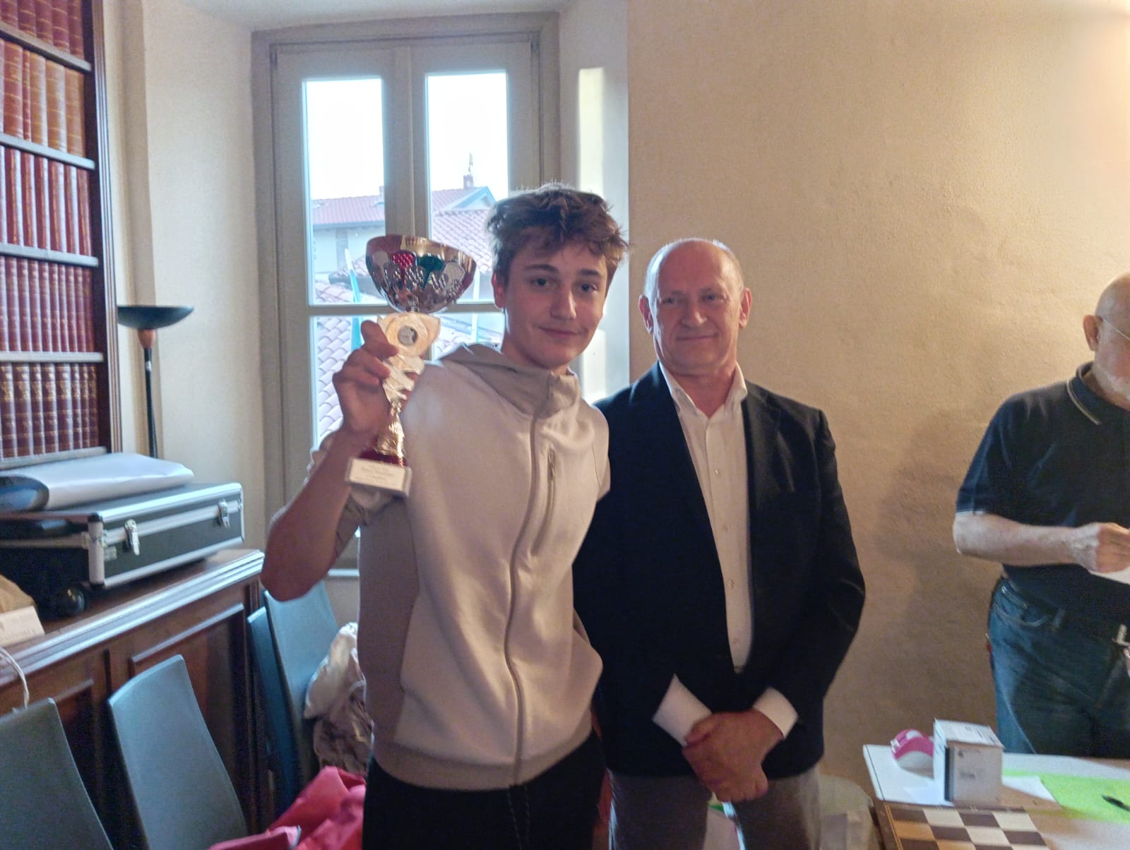 Un giovane vincitore di un torneo di scacchi tiene in mano una coppa, mentre sorride accanto a un uomo adulto, in una sala con libri e tavoli da gioco.