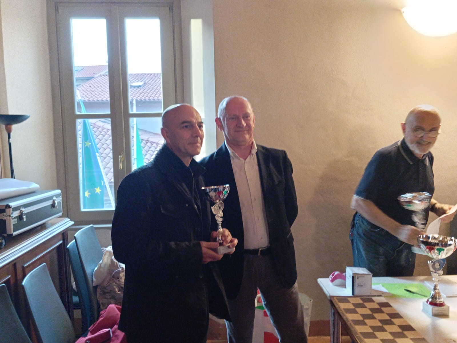 Due uomini sorridenti in una cerimonia di premiazione, uno dei quali tiene un trofeo, all'interno di una sala con finestre e bandiere visibili sullo sfondo.