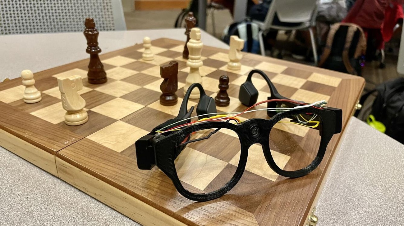 Occhiali tecnologici posizionati accanto a una scacchiera, con pezzi bianchi e neri in gioco.