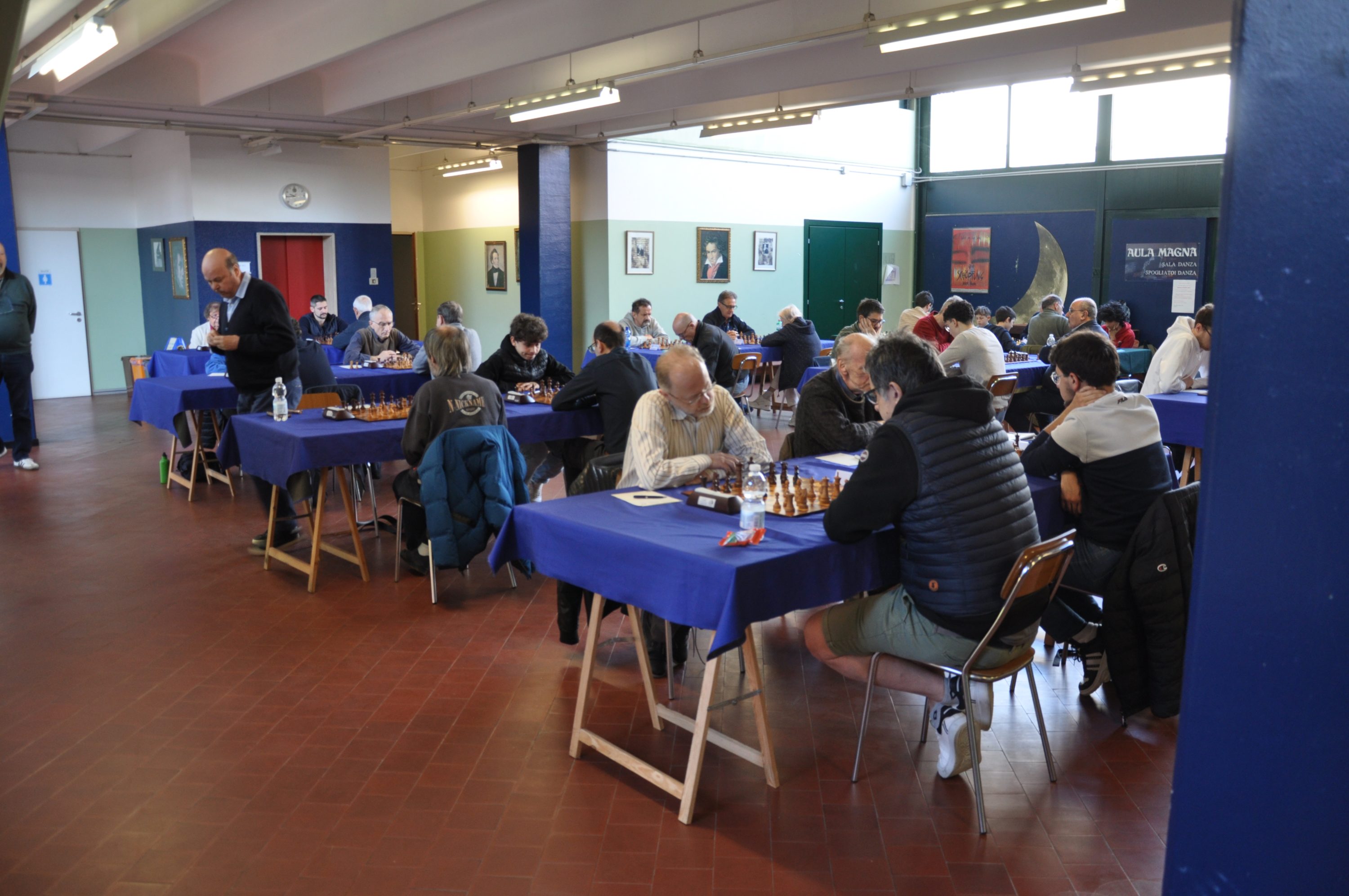 A chess tournament taking place indoors with multiple tables set up, each occupied by players engaged in matches. The room features blue tablecloths, and participants are focused on their games.