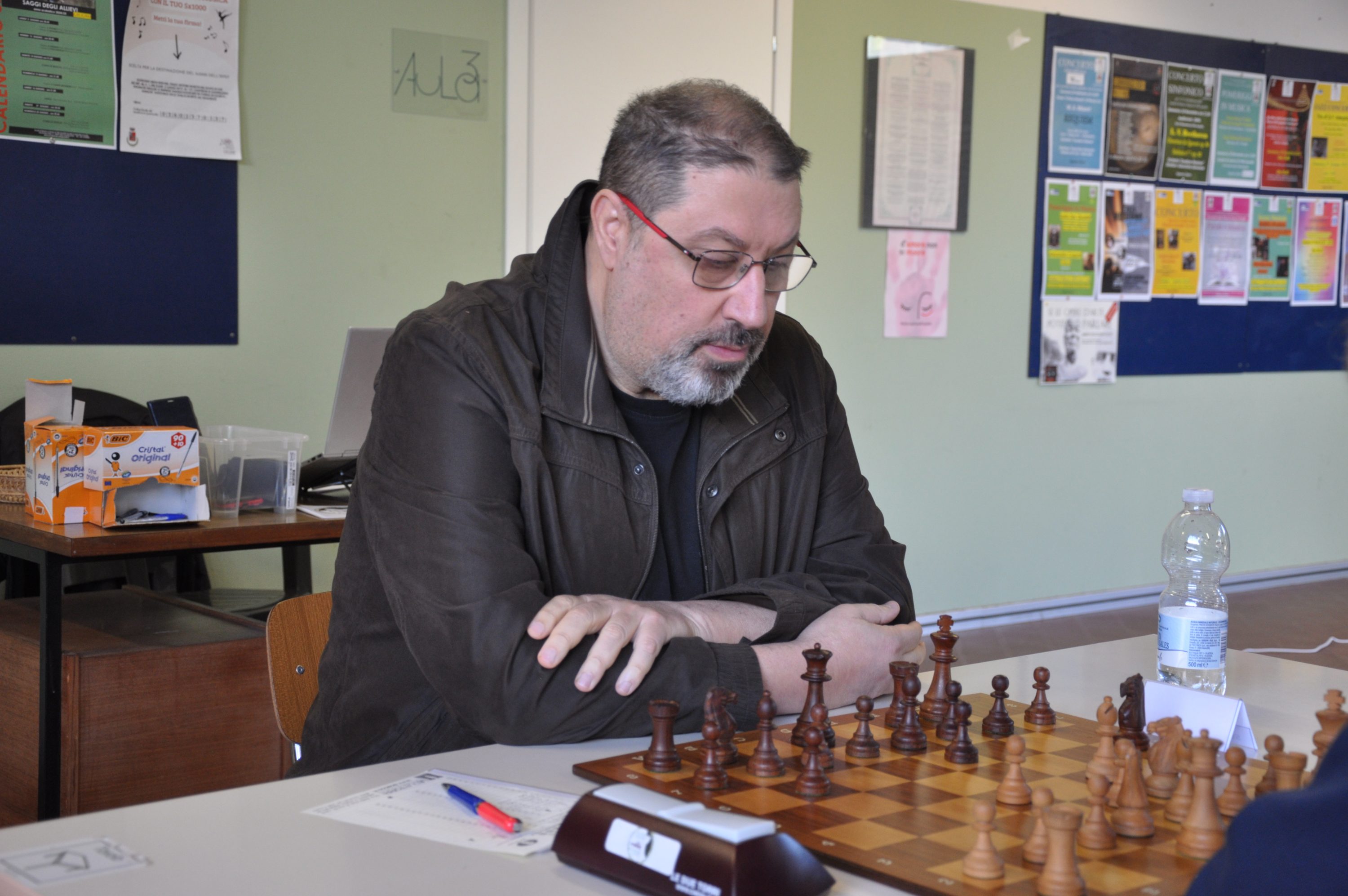A person deeply focused on a chess game, sitting at a table with a chessboard and pieces in view. The background shows wall posters and some items on a nearby table.
