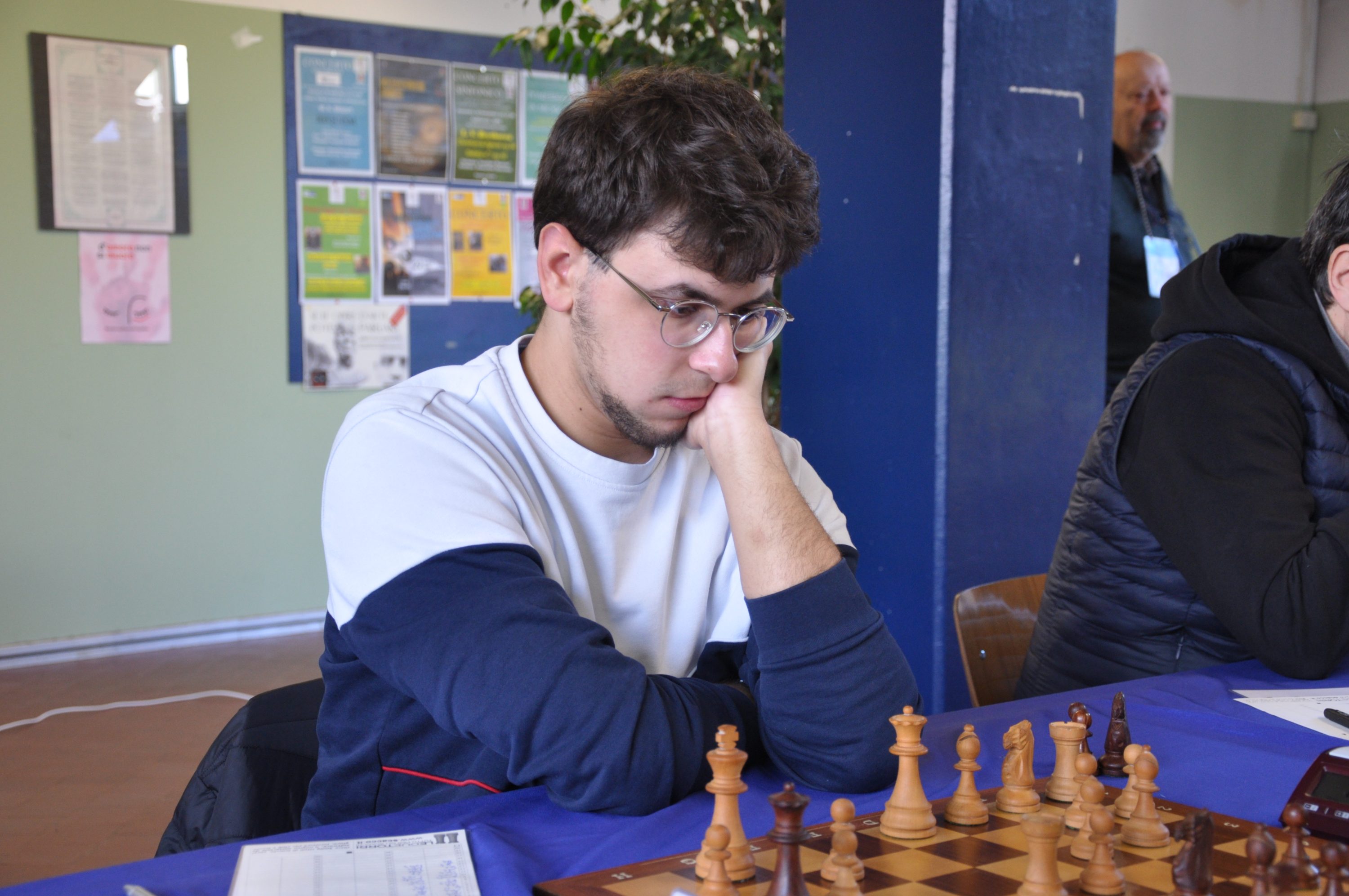 A young man with glasses is intently thinking while playing chess at a tournament, with a chessboard and pieces in front of him.