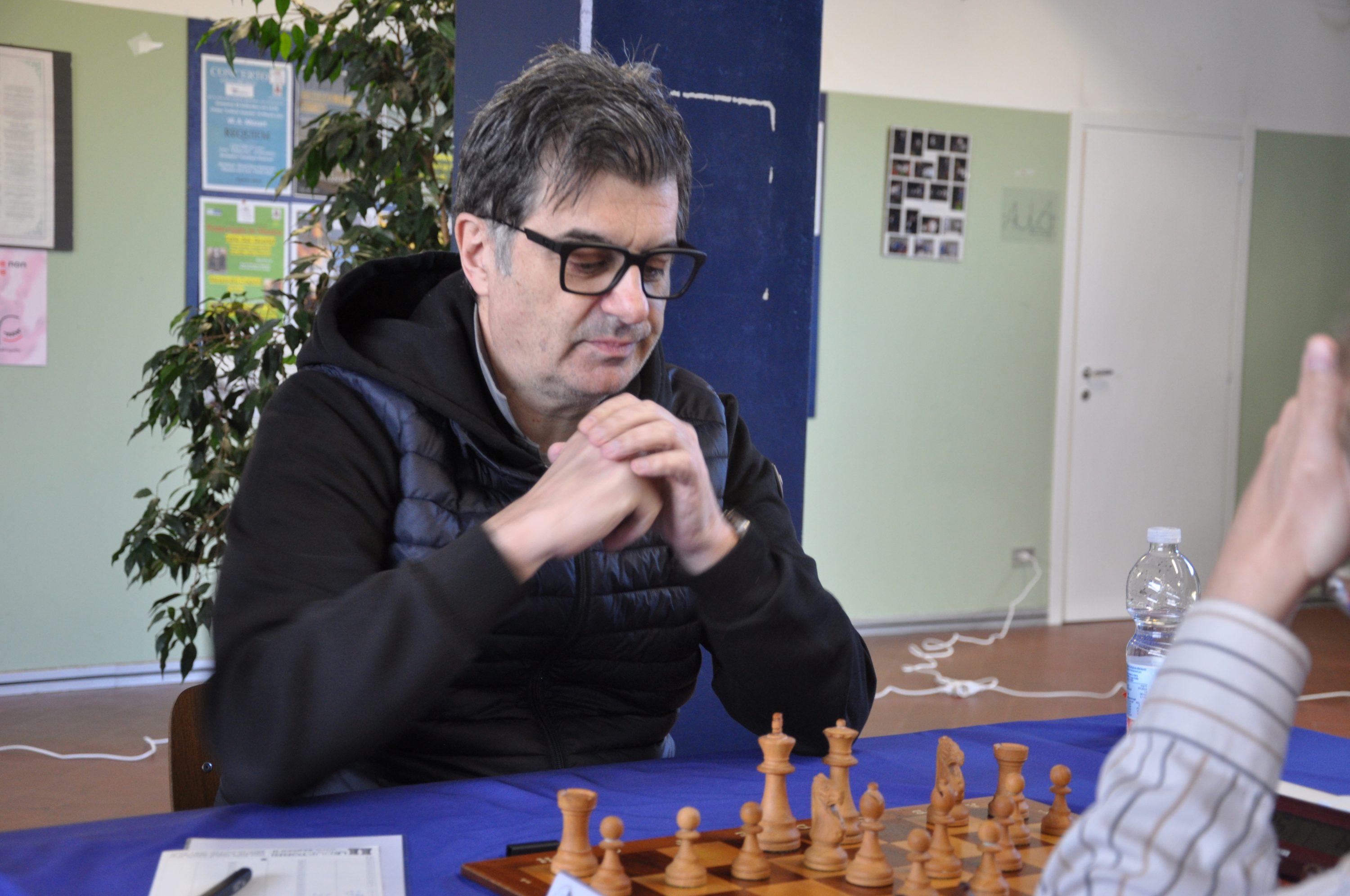 A thoughtful man in glasses sits at a chessboard, pondering his next move, with a blurred hand of another player visible across the table.