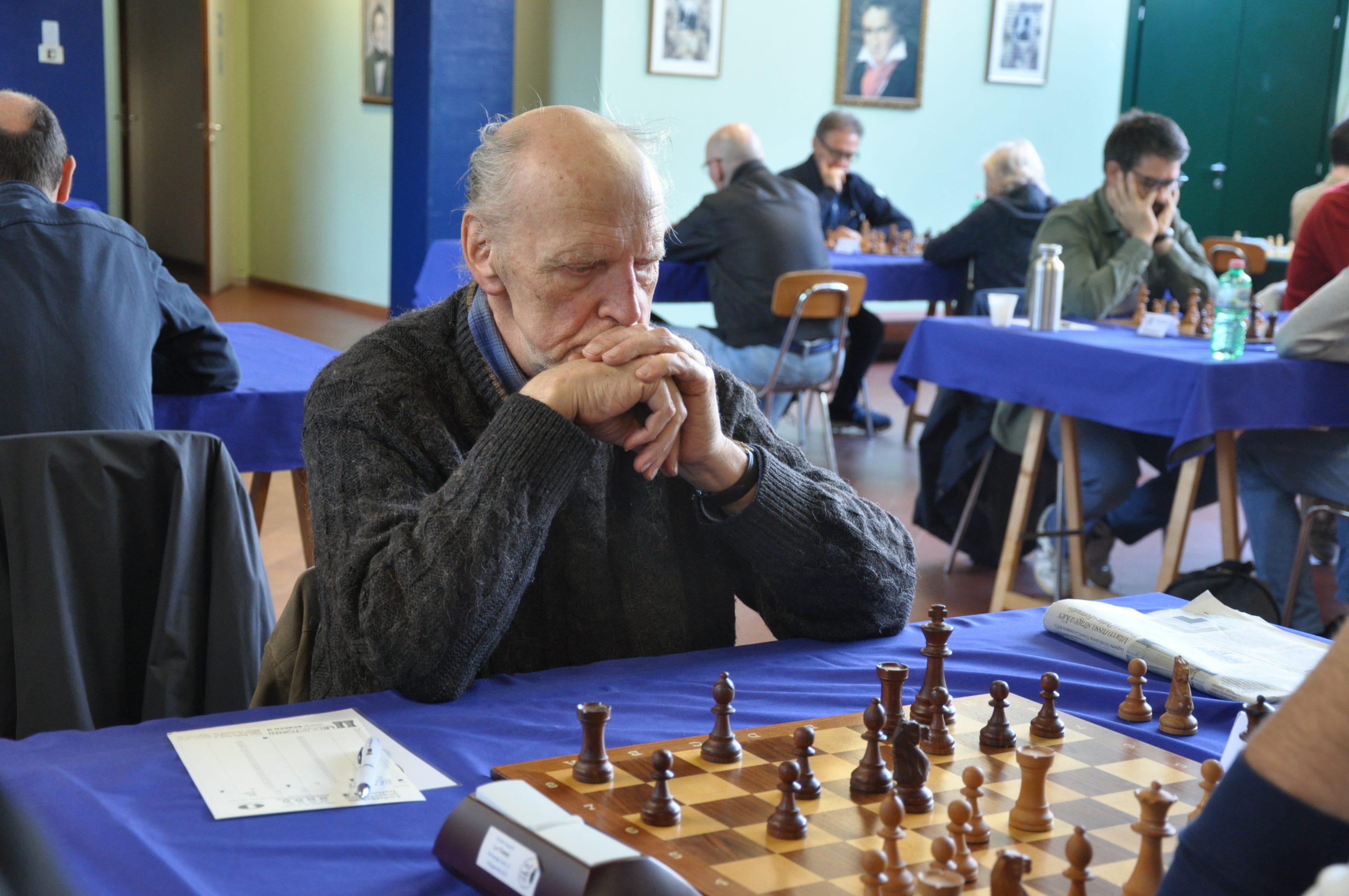 An elderly man deep in thought while playing chess at a tournament, surrounded by other players at nearby tables.