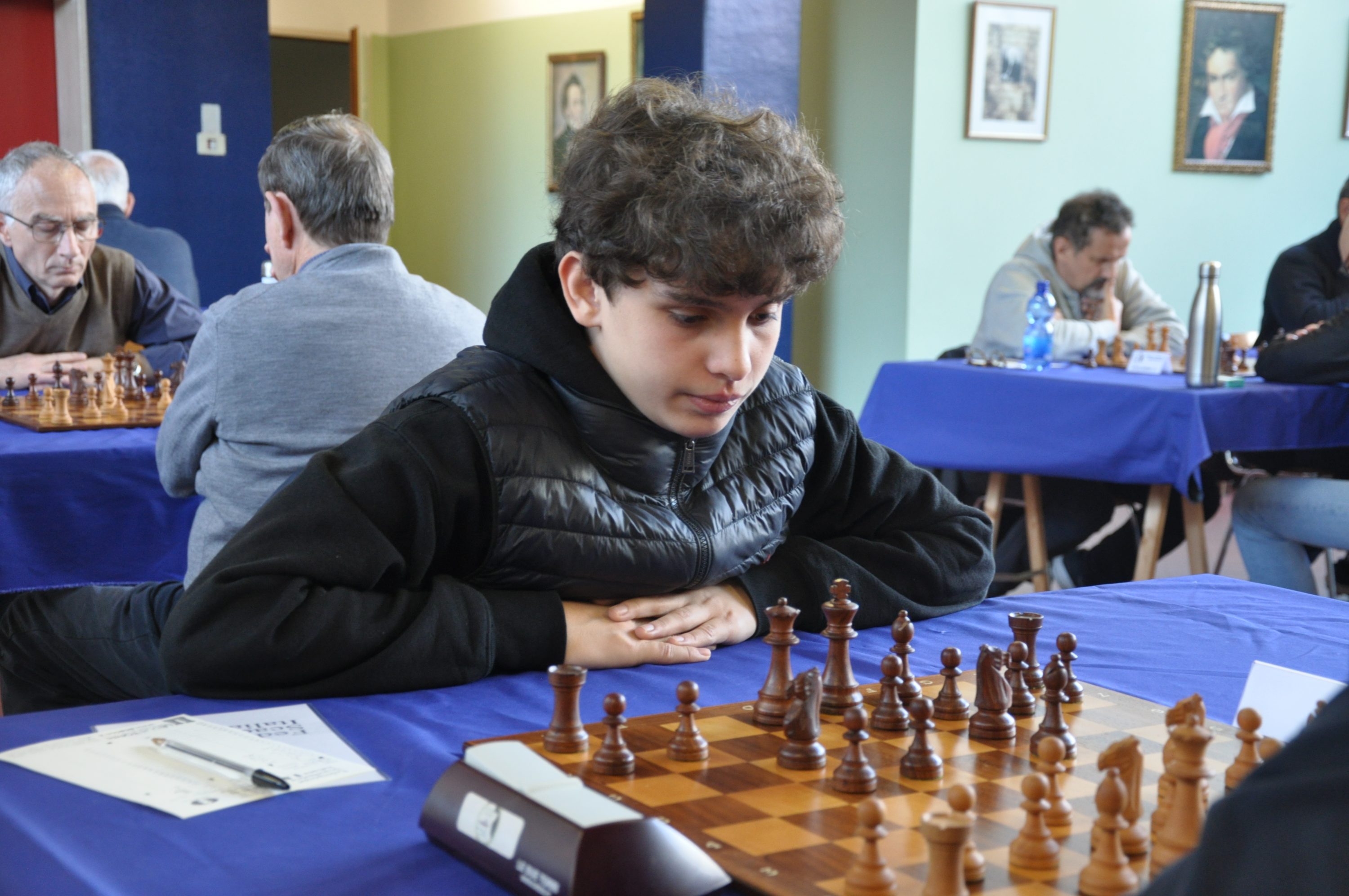 A young boy with curly hair, focused on a chess game, resting his chin on his hand, with other players visible in the background.