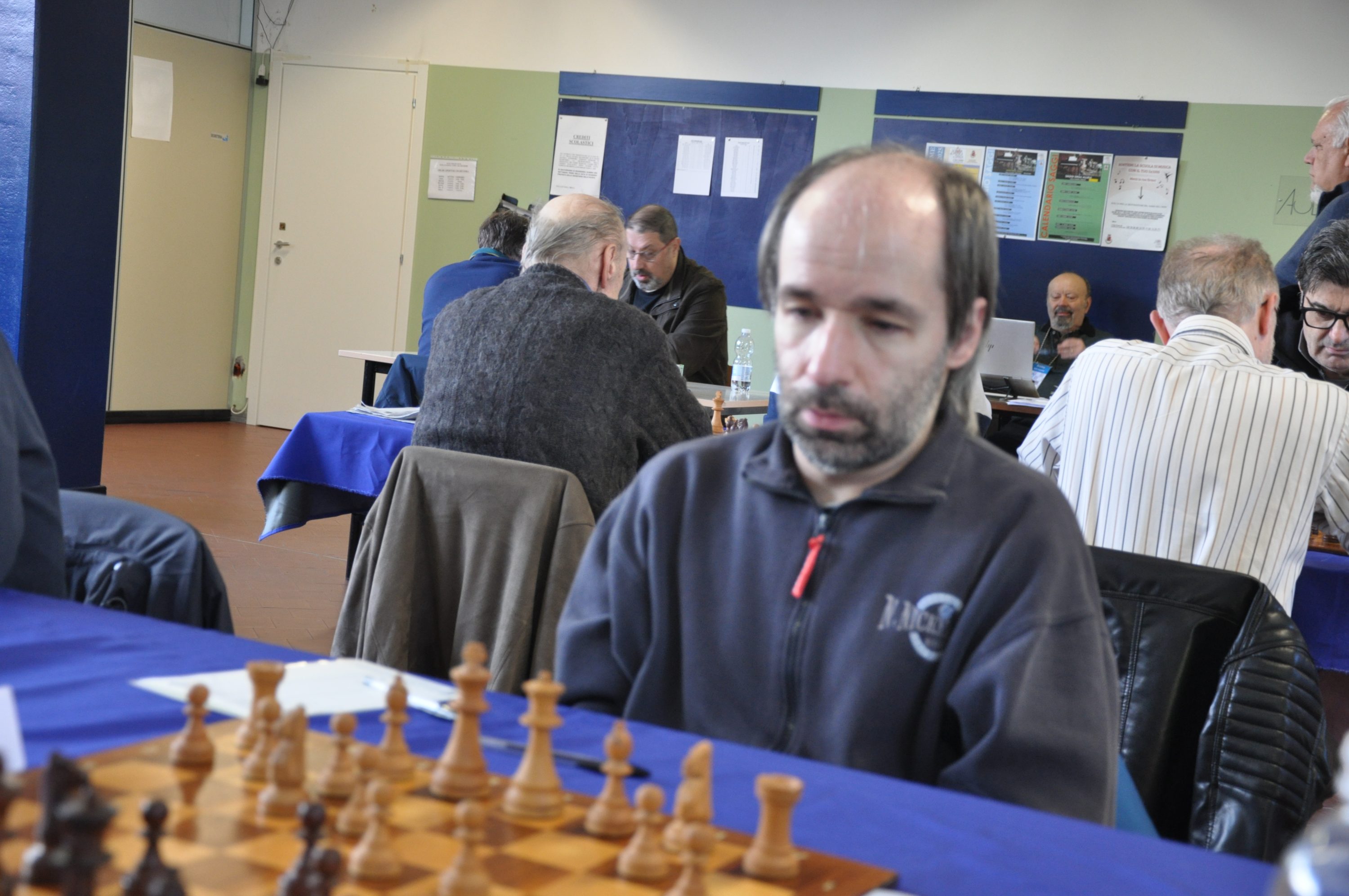 A focused player deep in thought, sitting at a chess table with a chessboard and pieces visible in the foreground. Other players and tables are seen in the background, creating a lively chess tournament atmosphere.
