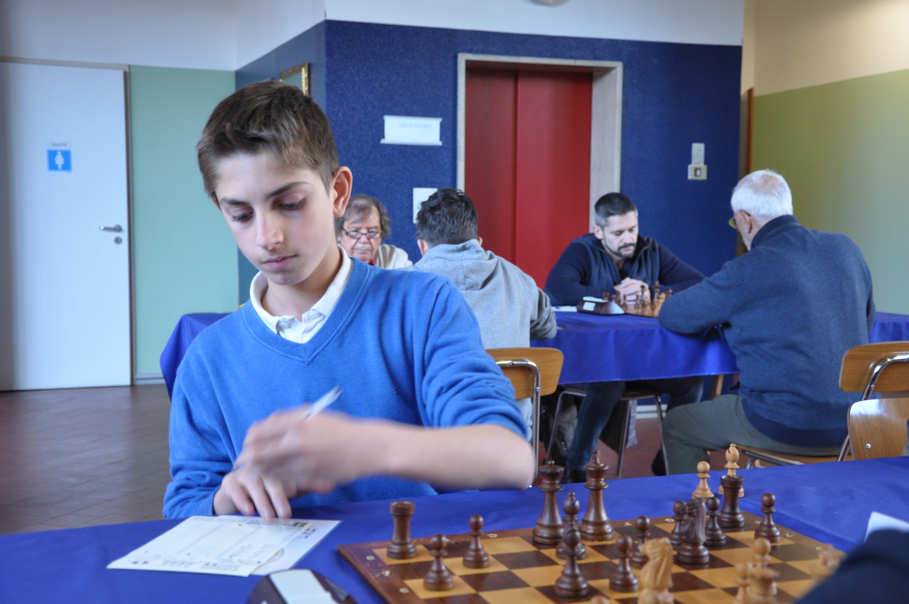A young boy in a blue sweater is sitting at a chess table, focused on writing notes while a chessboard with pieces is in front of him. Other players can be seen in the background, engaged in their games.