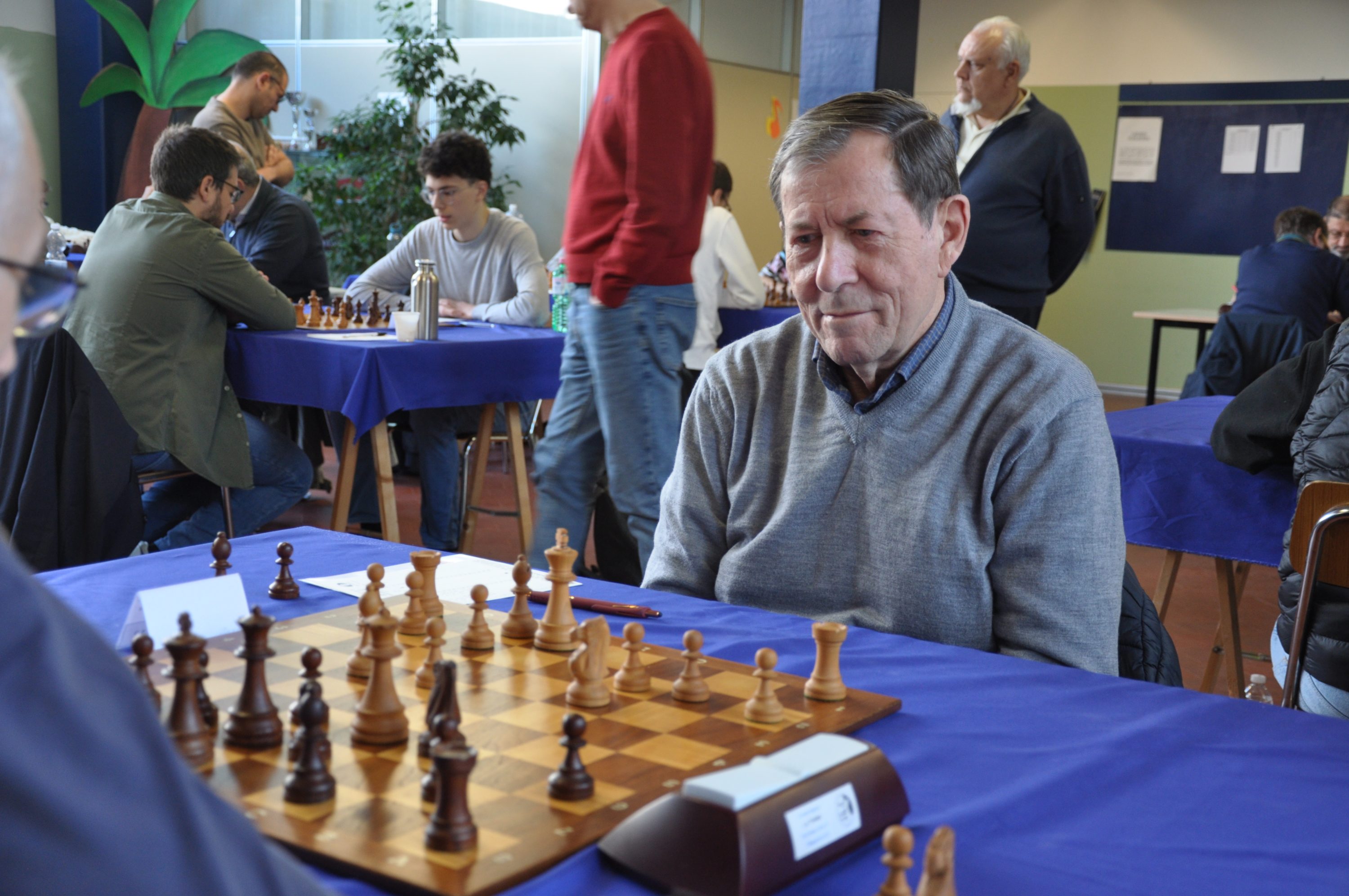 A chess player deeply focused on a game, sitting at a table with a chessboard and pieces. Other players and spectators are visible in the background.