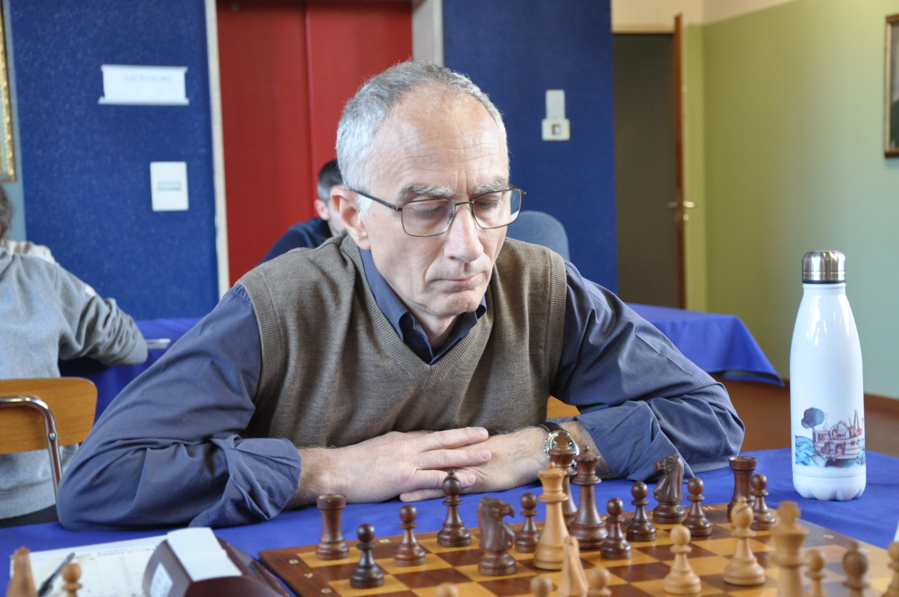 An elderly man concentrating on a chess game, seated at a table with a chessboard and pieces in front of him.