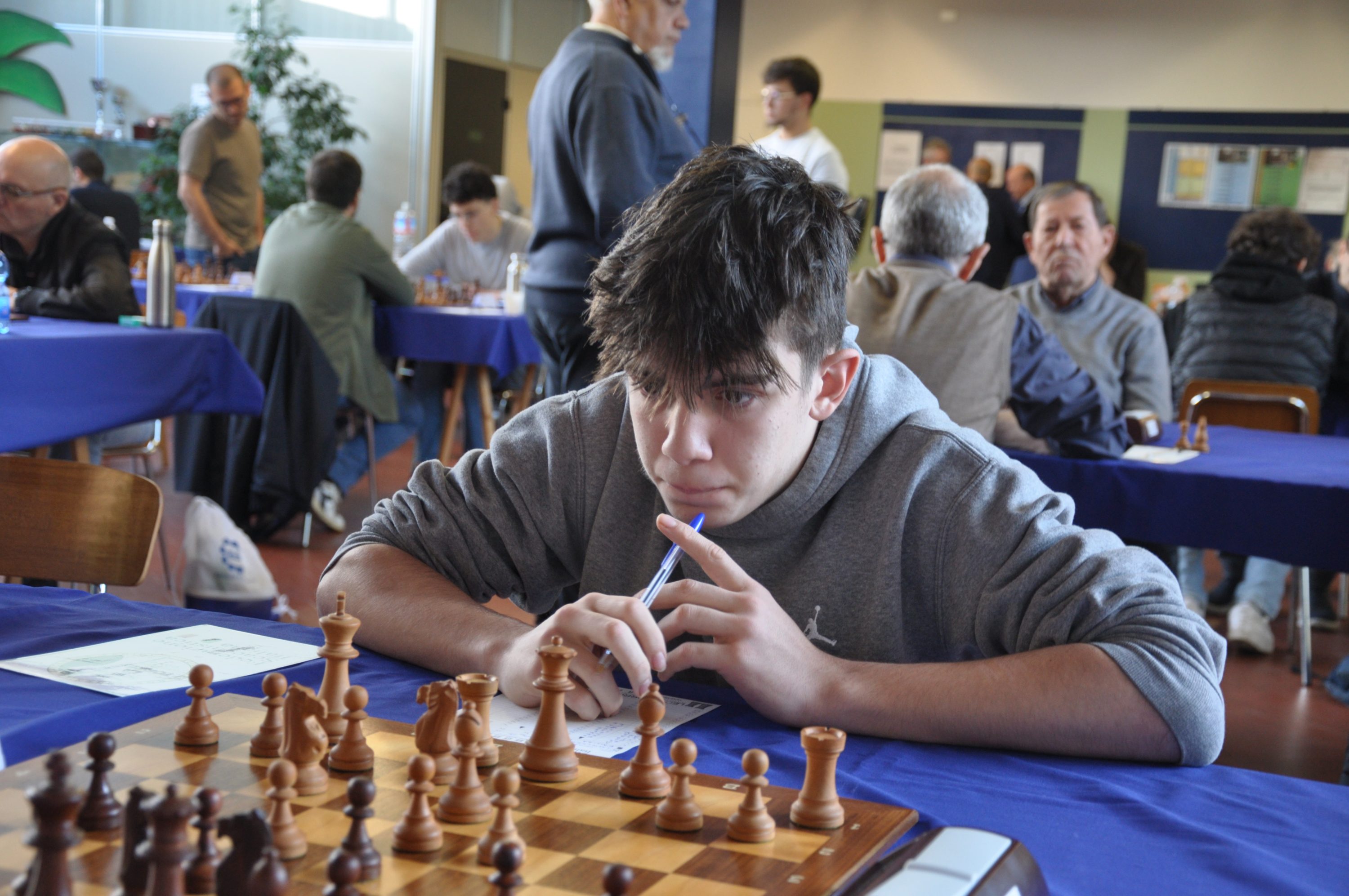 A young man deeply focused on a chess game at a competition, with a chessboard and pieces in front of him.