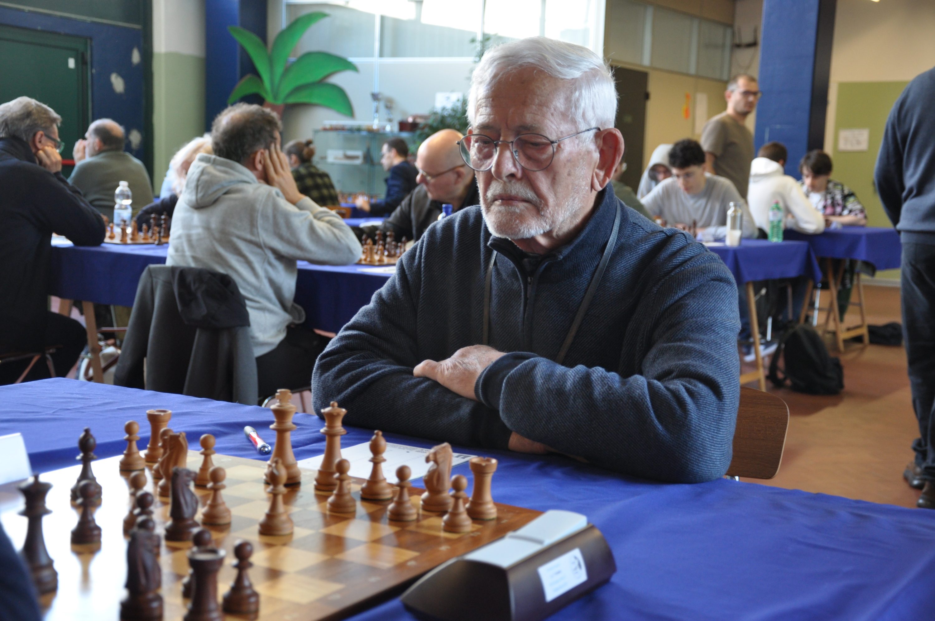 An elderly man with glasses is deeply focused on a chessboard during a tournament, with other players in the background.