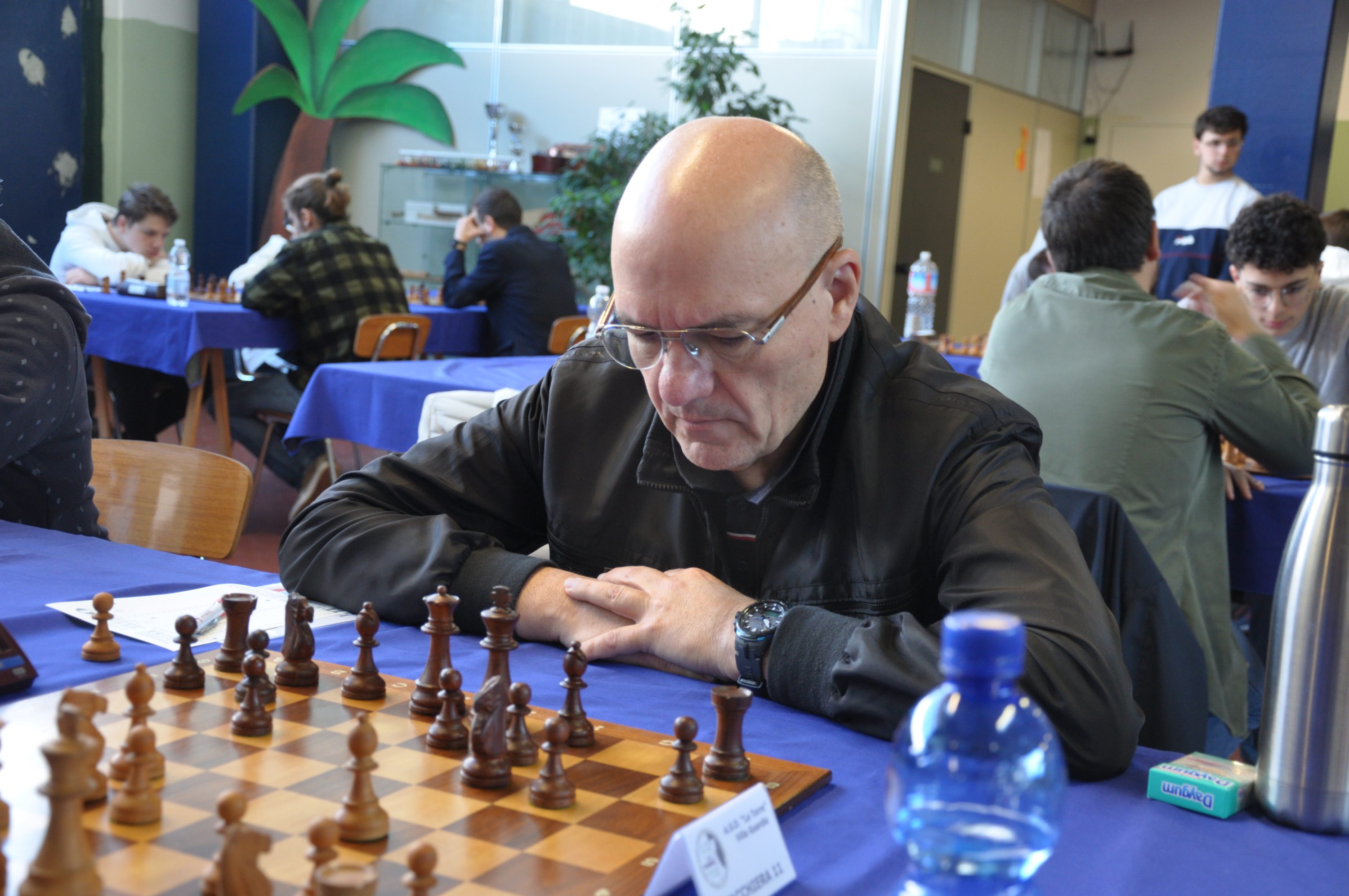 A focused chess player contemplating a move at a chess tournament, with a chessboard filled with pieces in front of him.