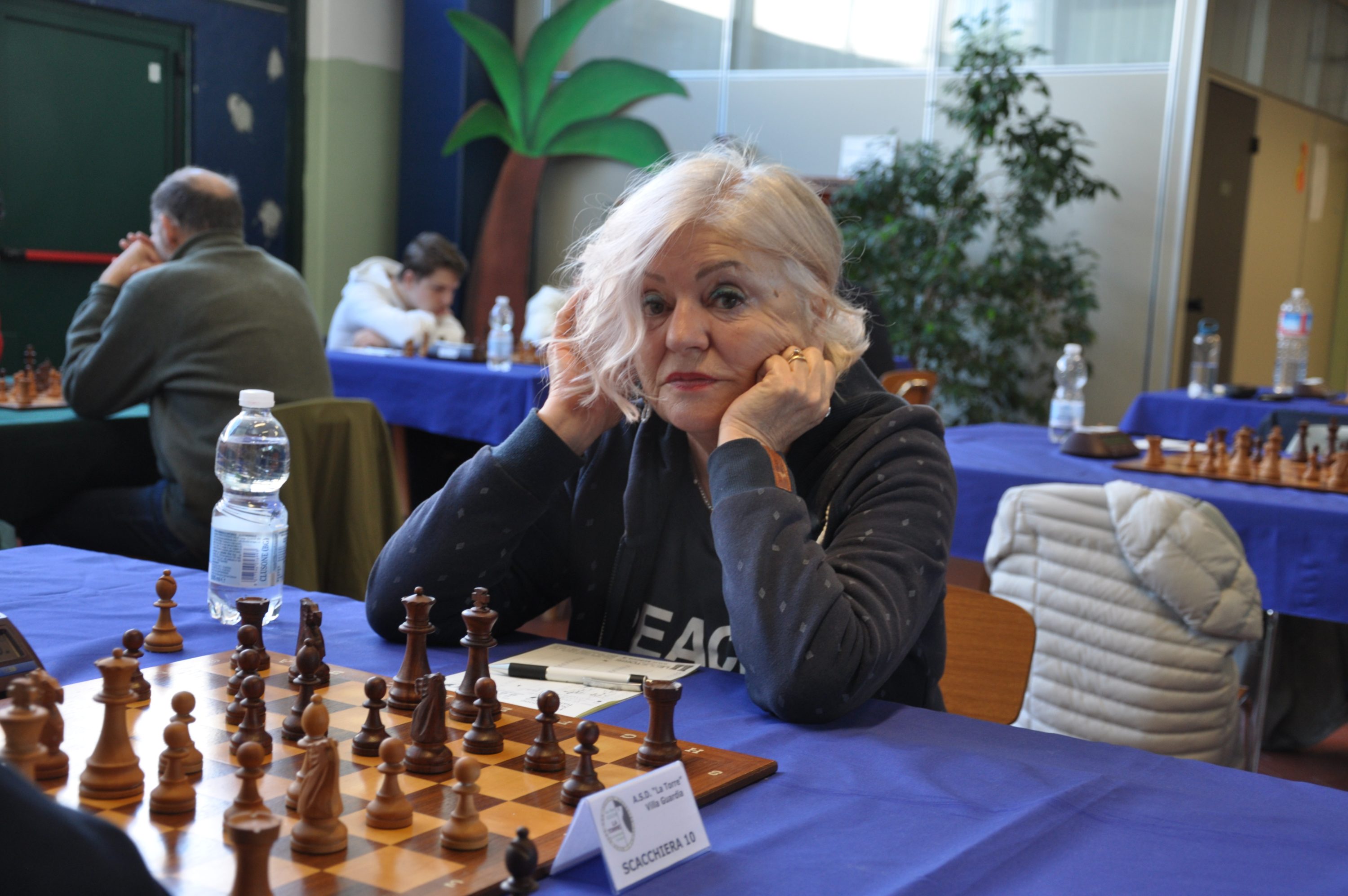 A focused chess player with short blonde hair sitting at a chessboard in a tournament setting, pondering her next move.