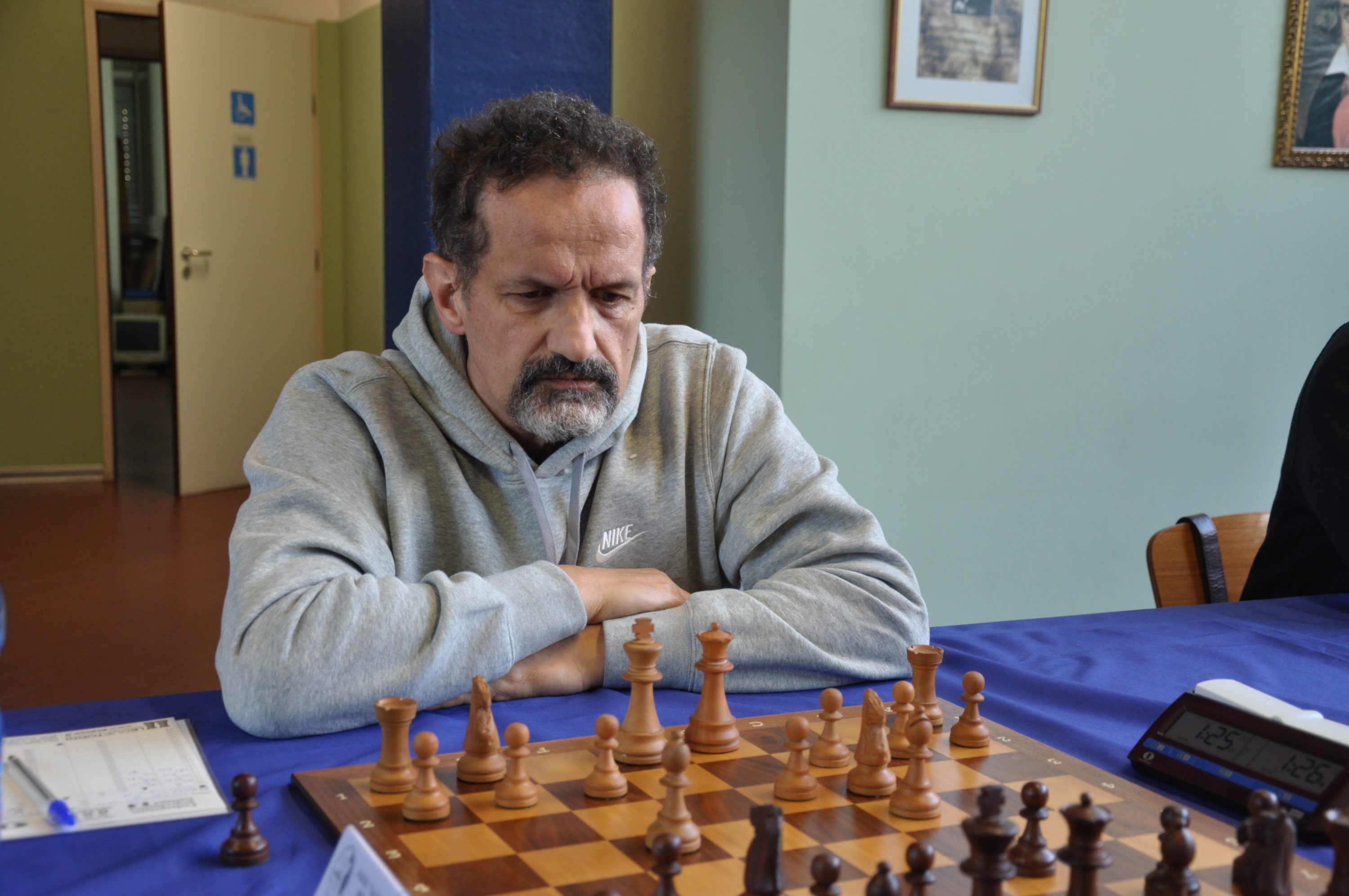 A man in a gray hoodie is concentrating on a chess game at a wooden chessboard with an assortment of pieces.