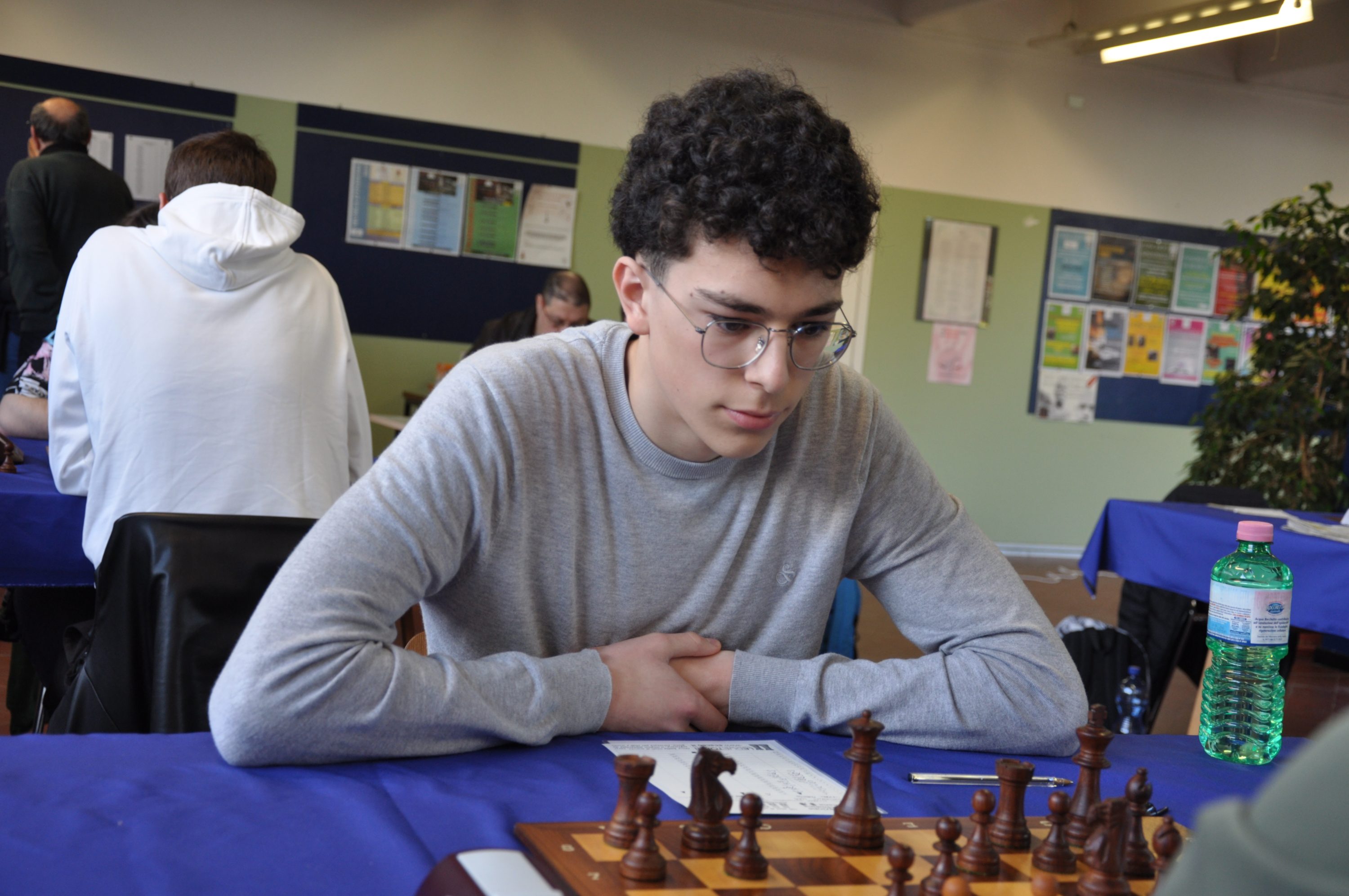A young man with curly hair and glasses concentrated on a chess game, seated at a table with chess pieces in front of him.