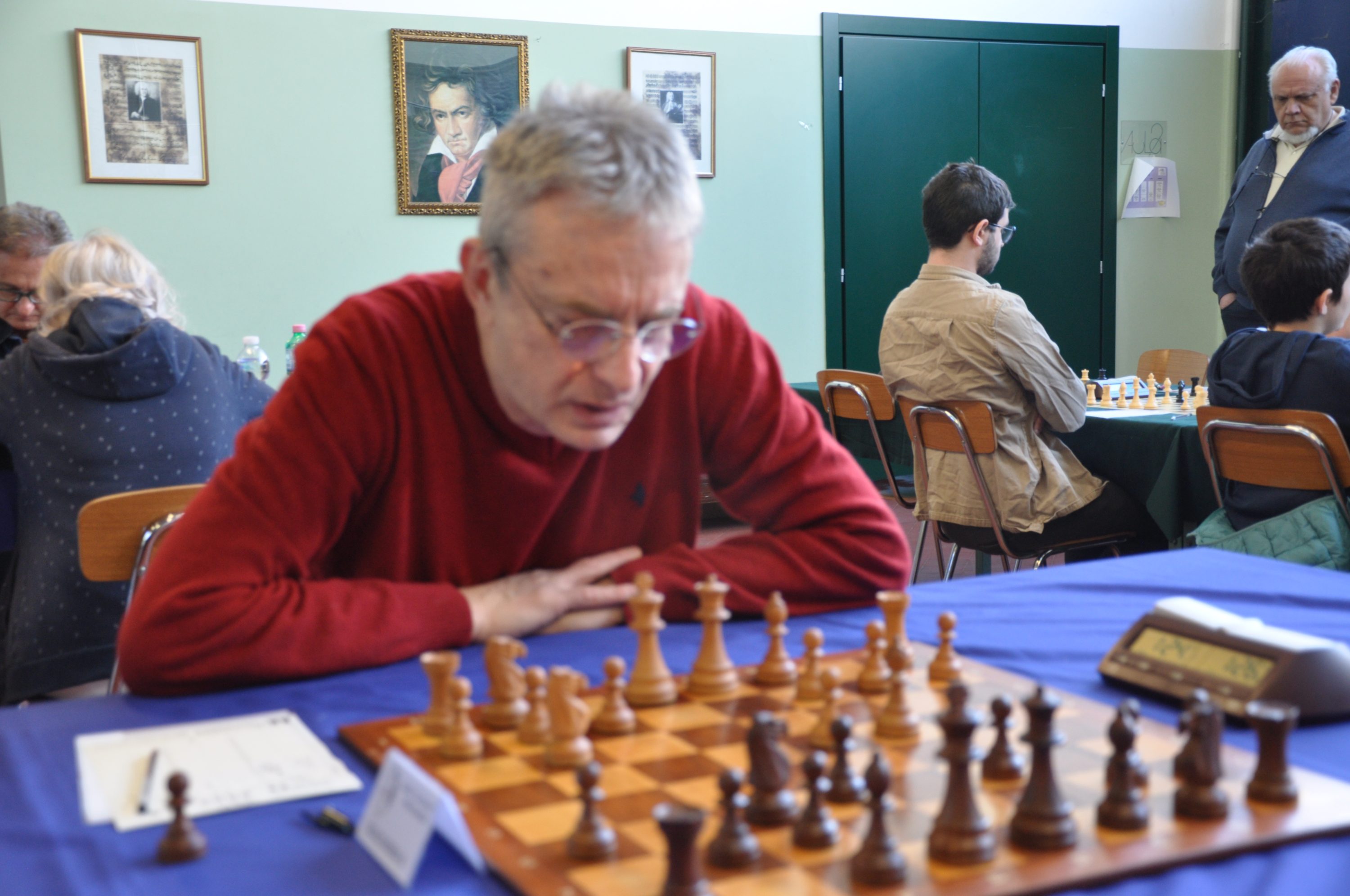 A focused individual in a red sweater is deeply engaged in a chess game, with a wooden chessboard and pieces in front of him. Other players can be seen in the background at a chess event.