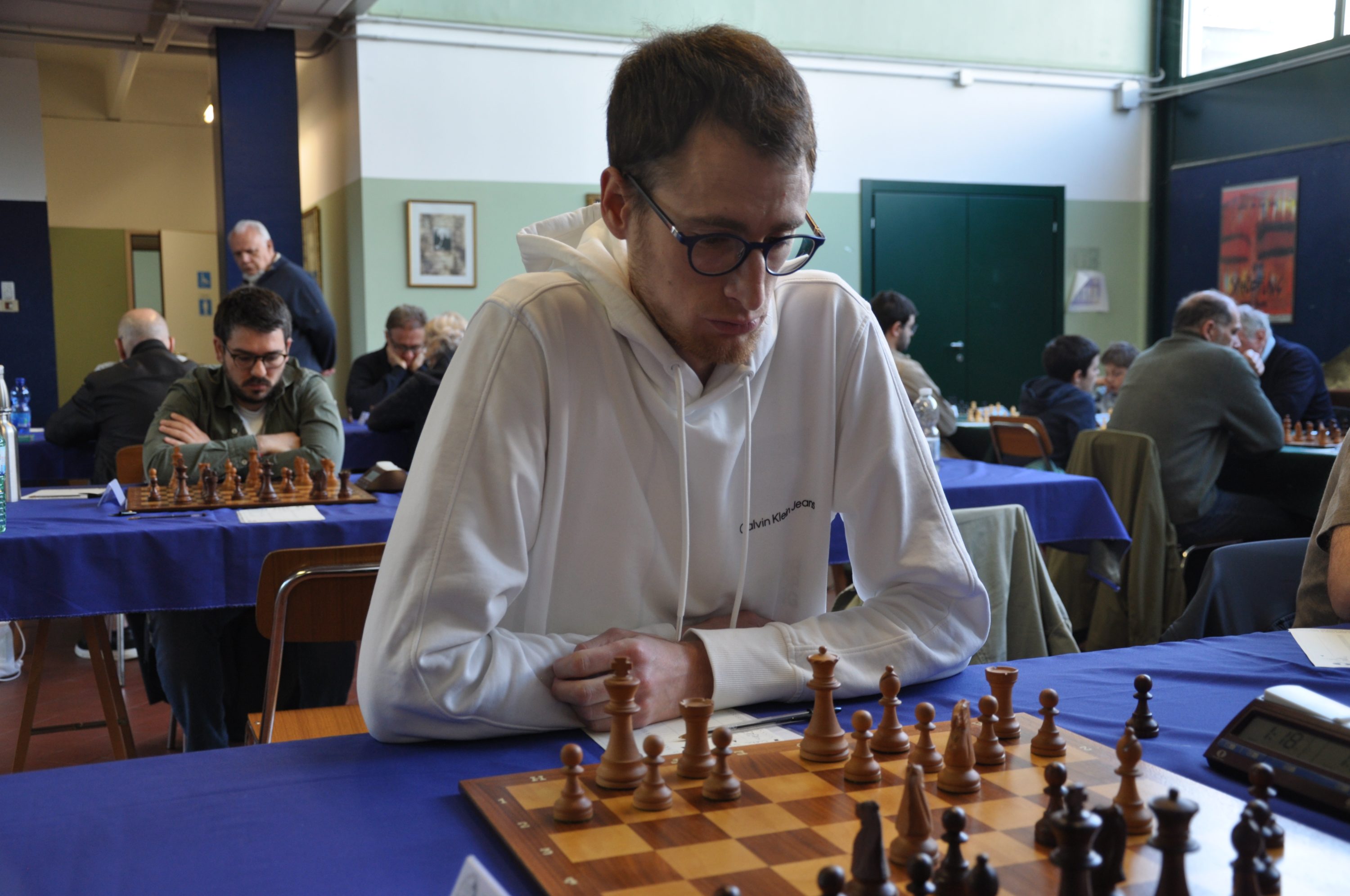 A chess player in a white hoodie focused on a chessboard during a tournament, with several other players and chessboards in the background.