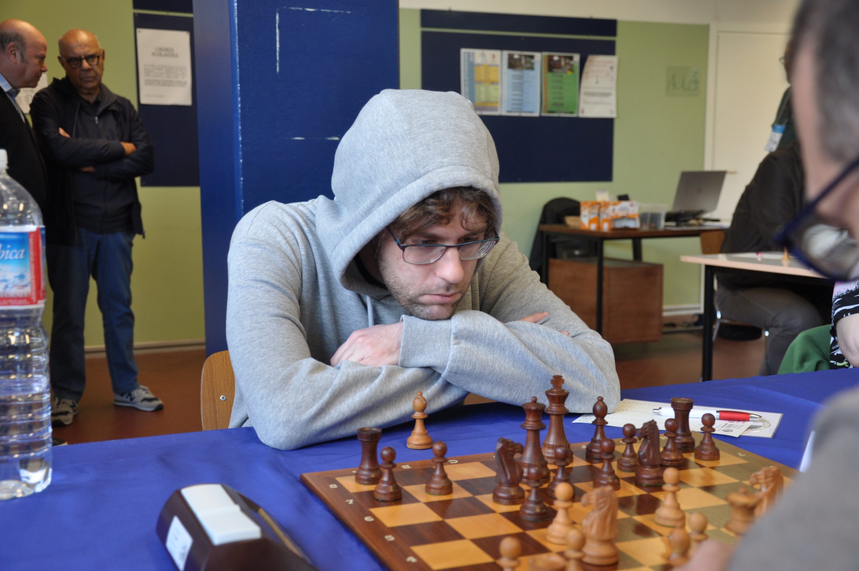 A person in a gray hoodie observing a chess game intently, with a chess board in front and spectators in the background.