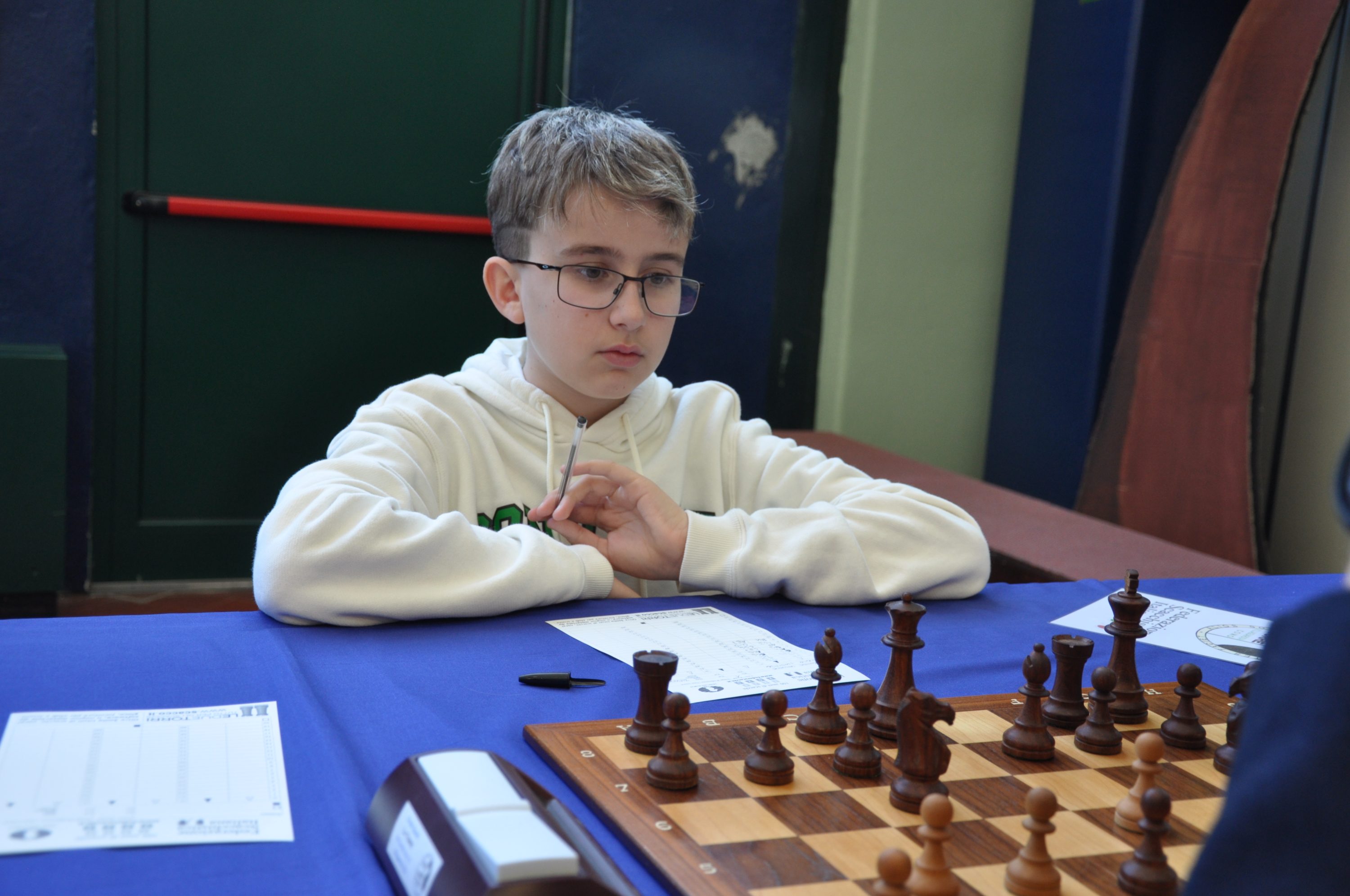A young player focused on a chess game, sitting at a table with a chessboard and score sheets.