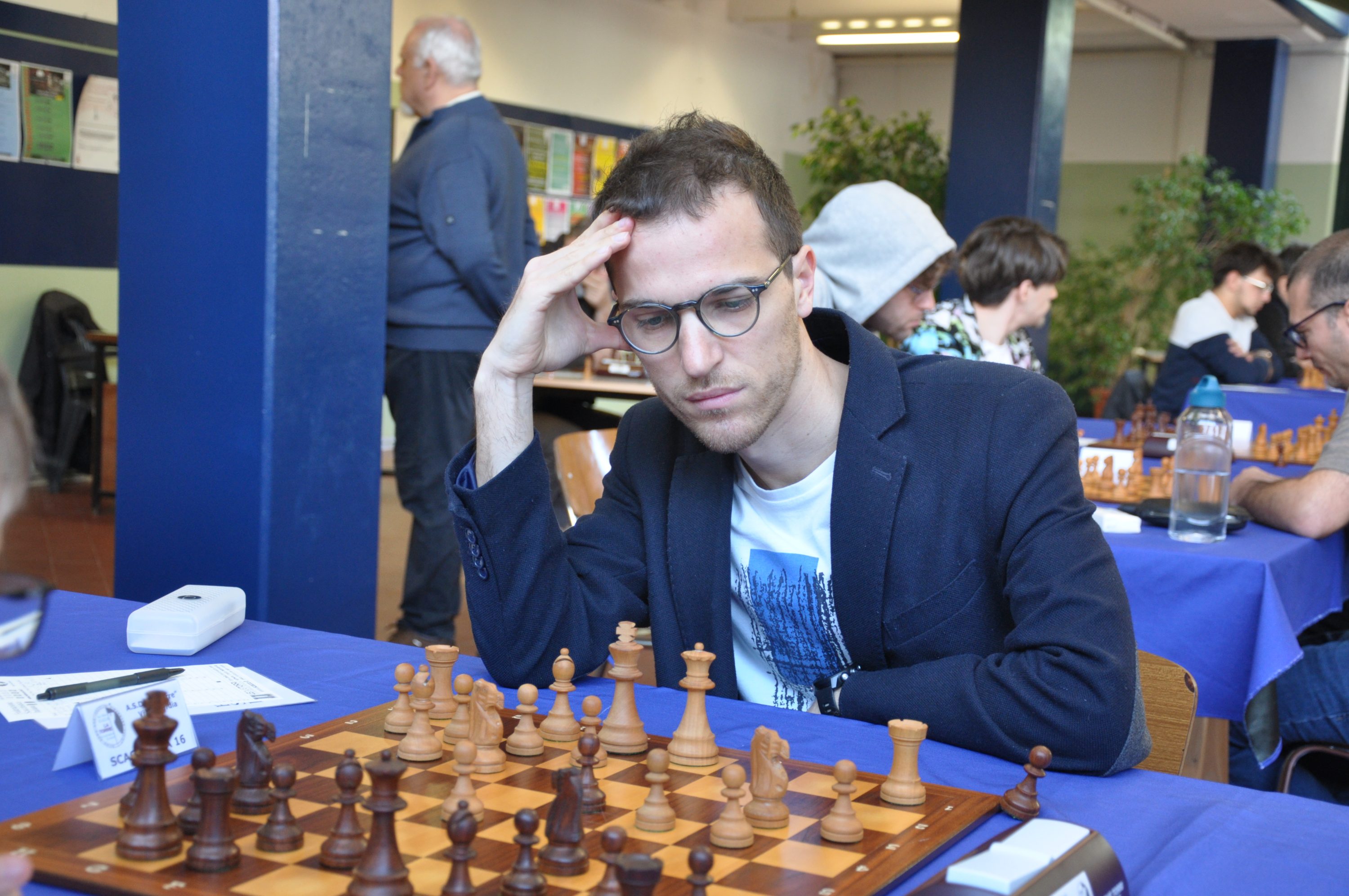 A focused chess player in a suit contemplates a move at a chessboard during a tournament.