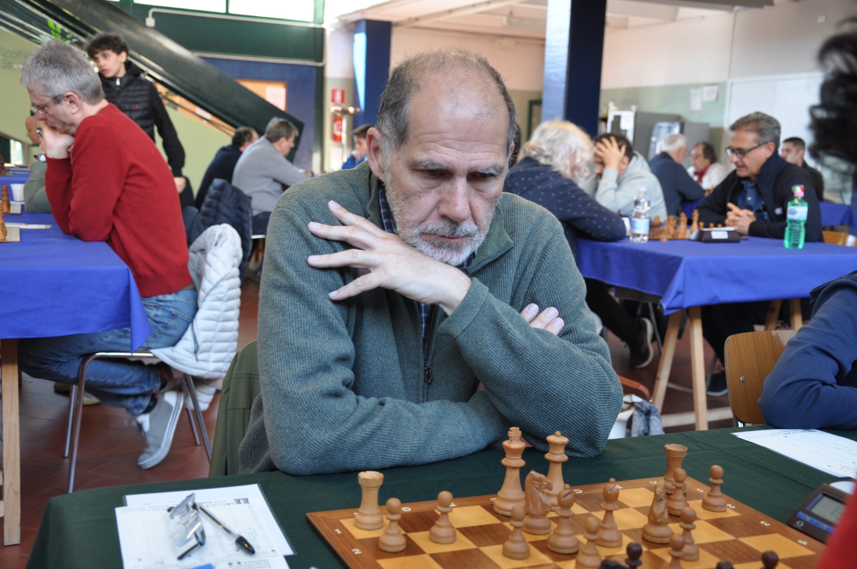 A man with a thoughtful expression is playing chess at a tournament, surrounded by other players and tables in a bright room.