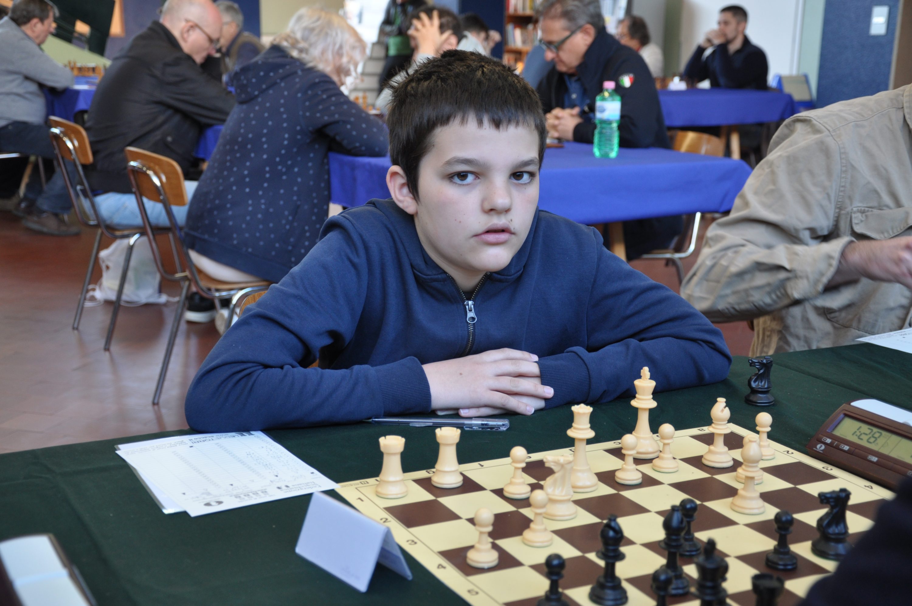 A focused young boy sitting at a chess table with his hands on the pieces, contemplating his next move in a chess tournament setting.
