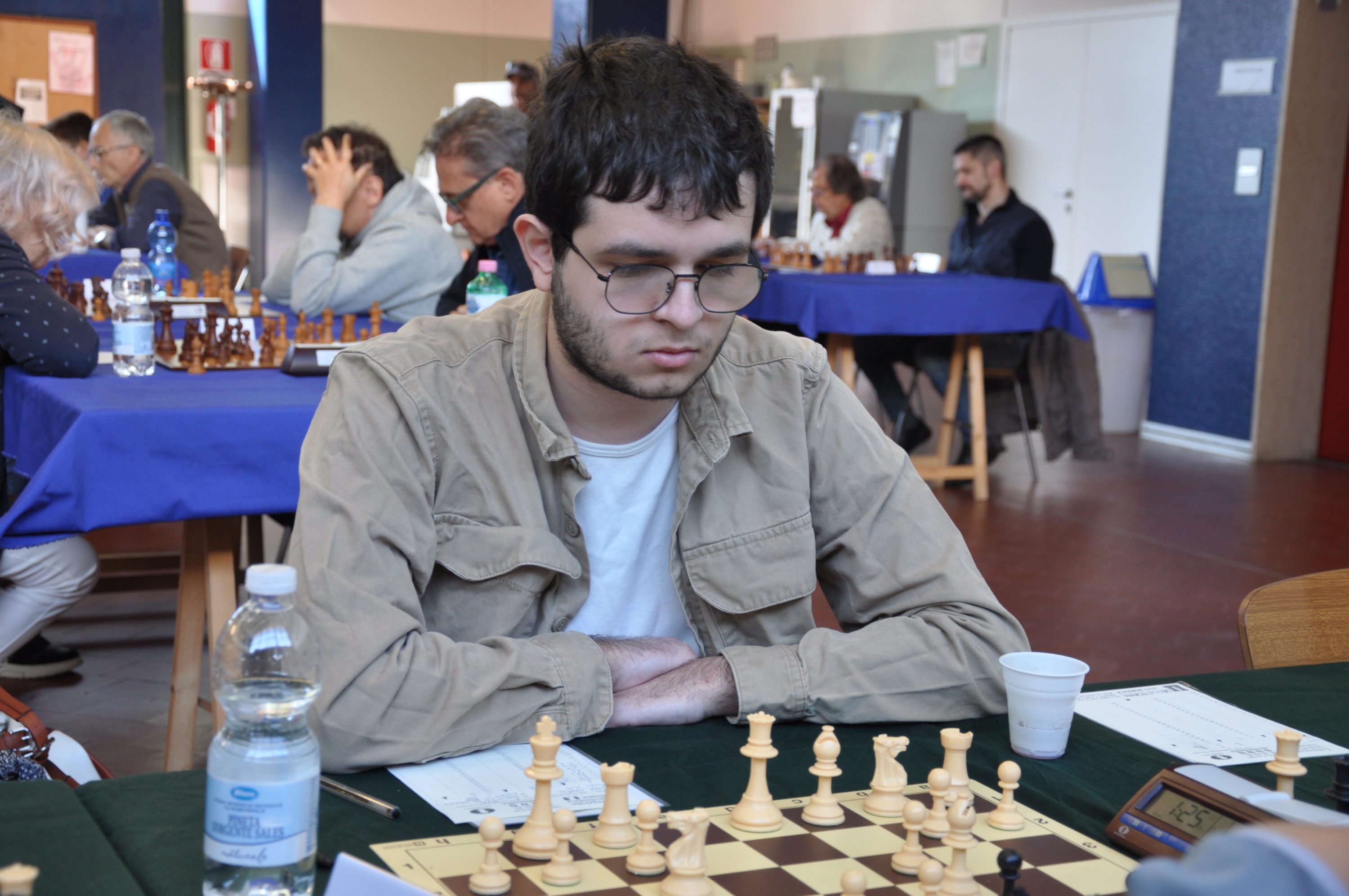A young man with glasses is focused on a chess game at a tournament, surrounded by other players in a well-lit room.