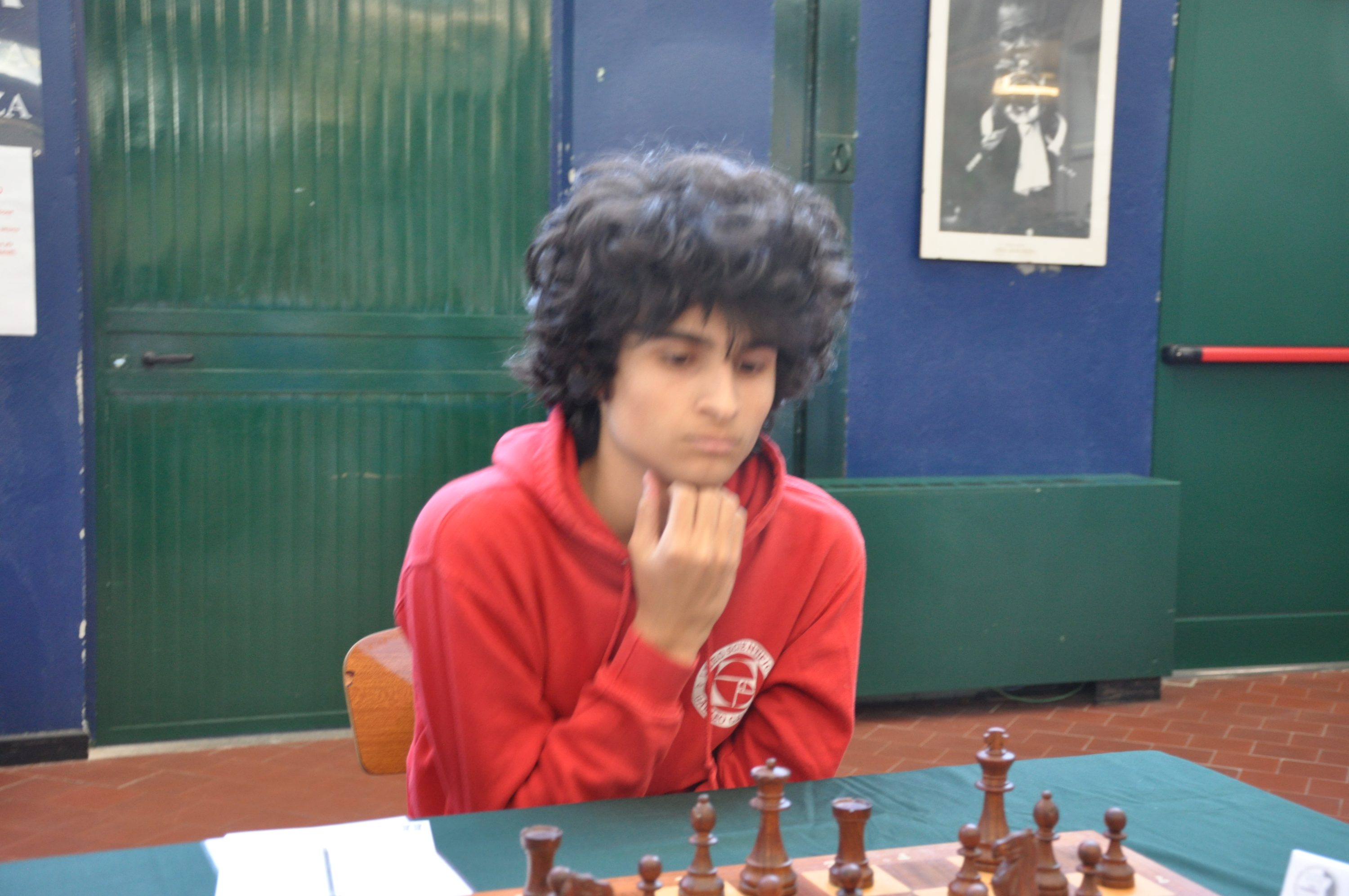 Young player with curly hair wearing a red hoodie, deep in thought while sitting at a chessboard.
