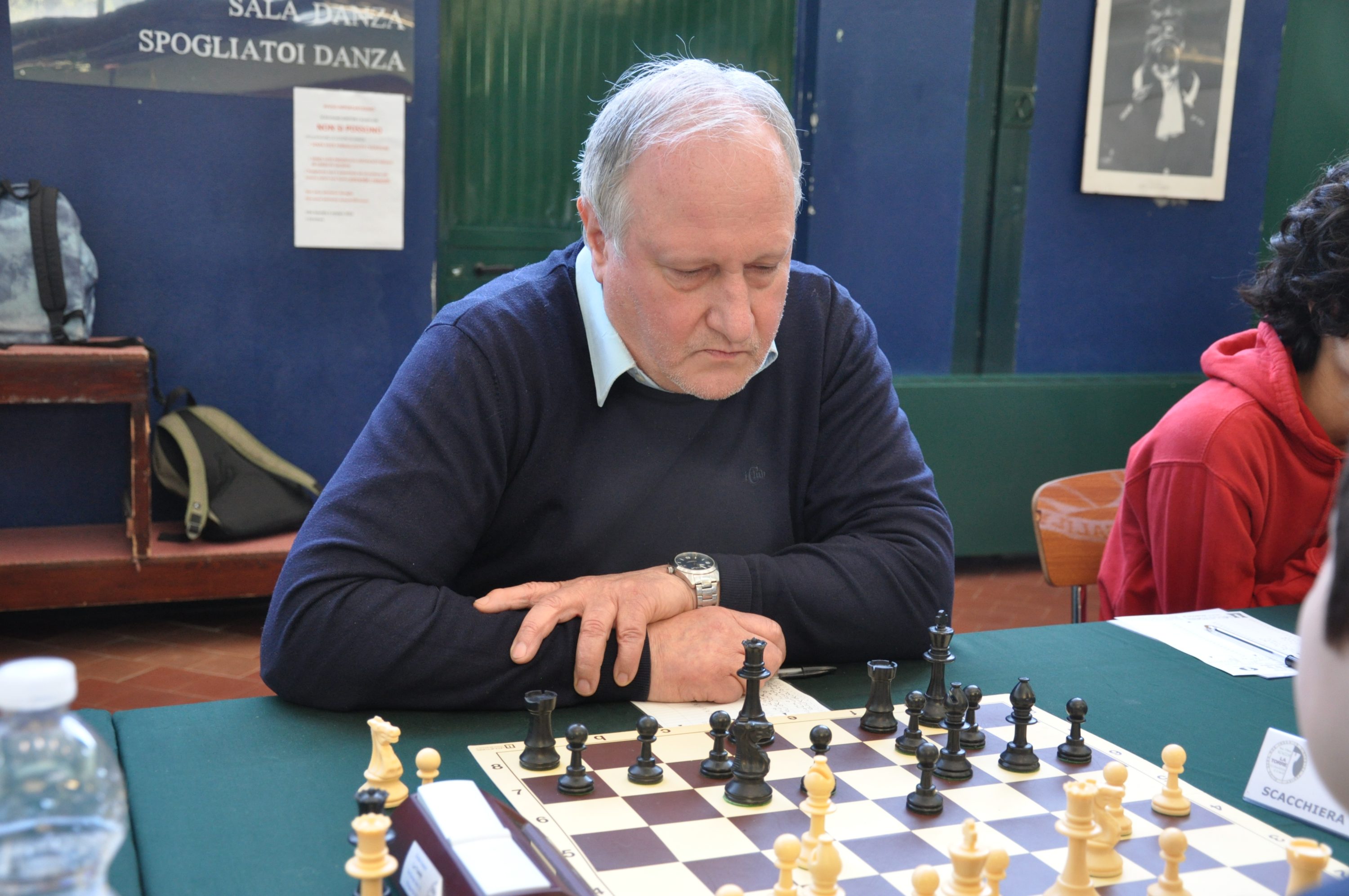 An older man with gray hair, wearing a dark sweater and shirt, is focused on a chess game with black and white pieces on a chessboard.
