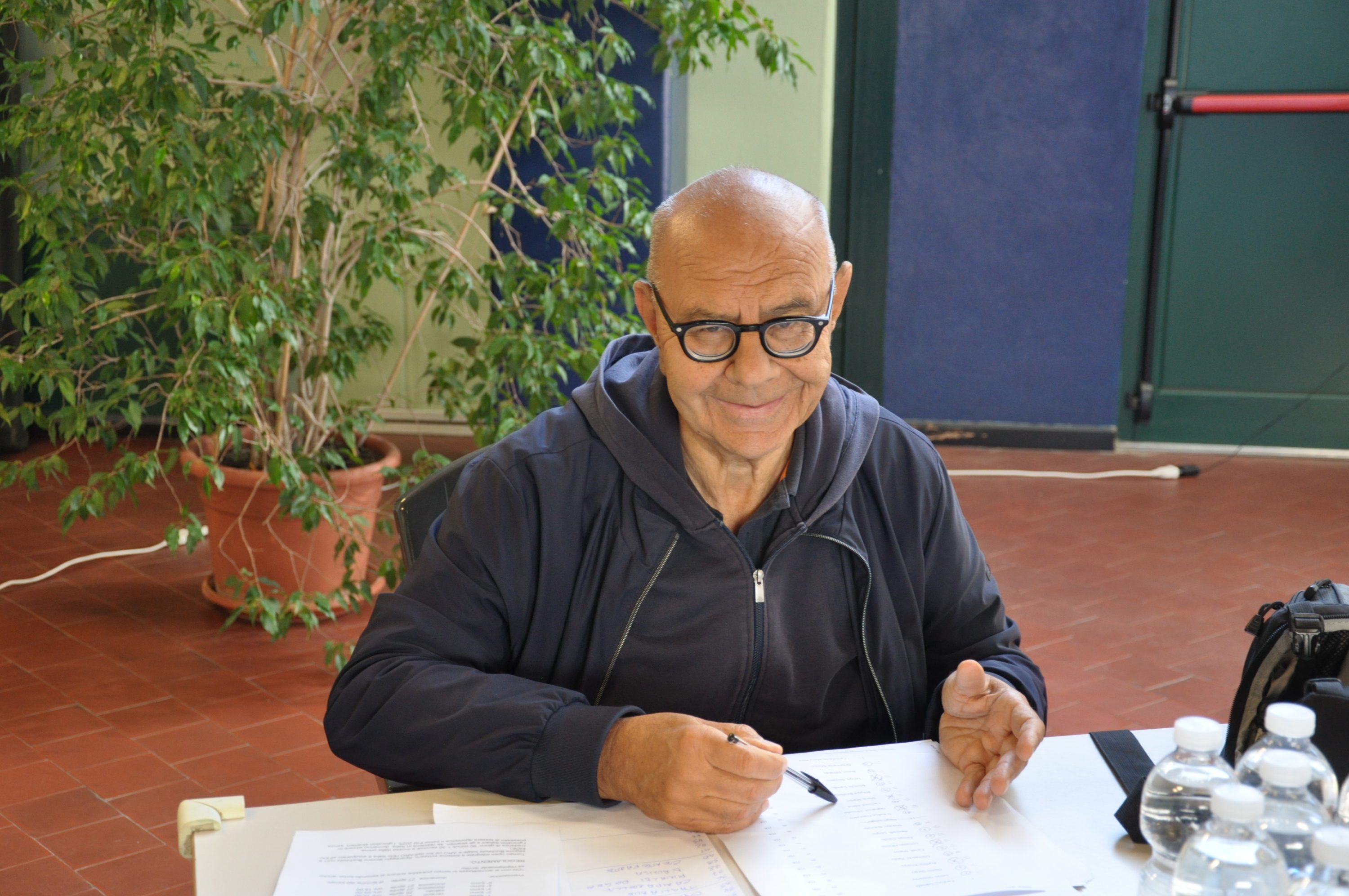 A smiling person wearing glasses sits at a table, with a plant in the background, engaged in writing or reviewing papers.