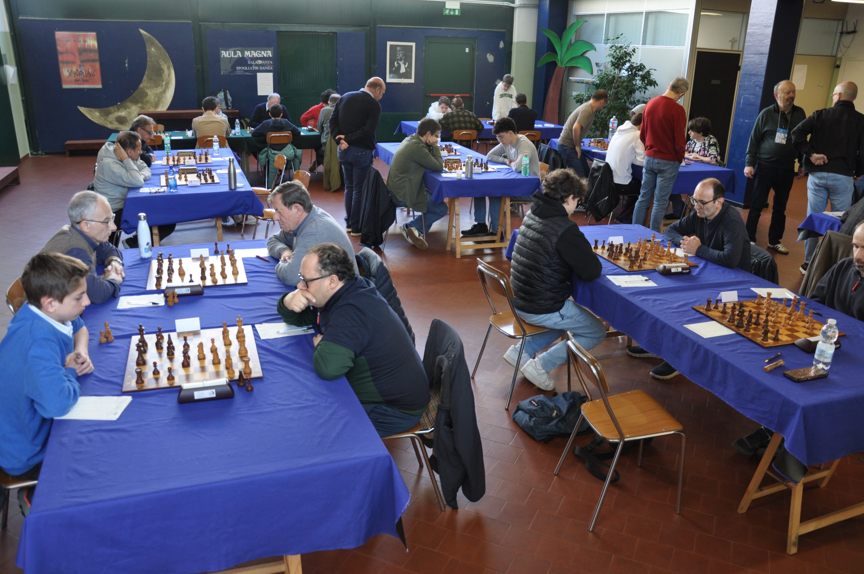 A chess tournament scene featuring several tables with players engaged in matches, all set against a background of blue tablecloths and a spacious indoor venue.