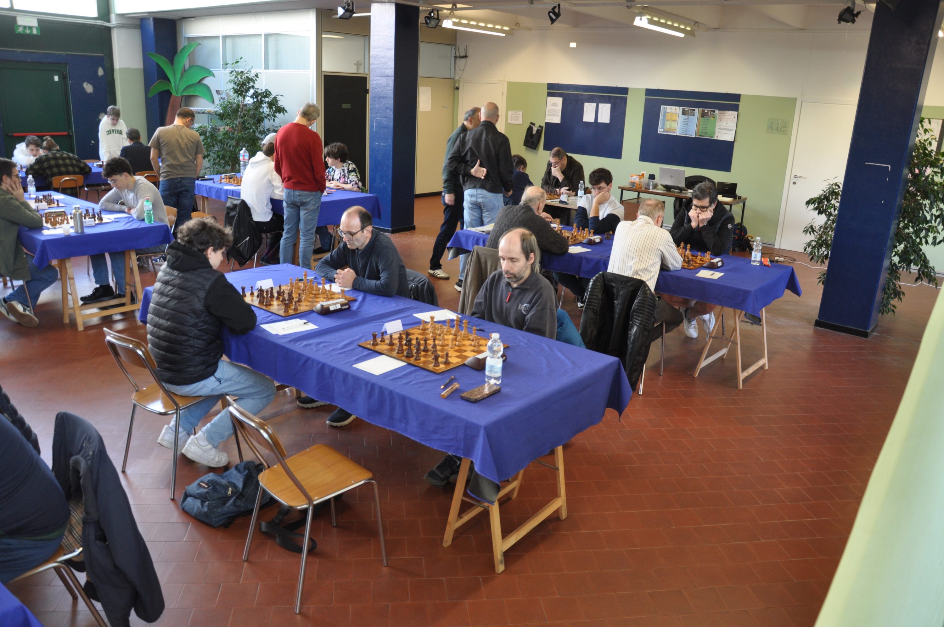 A chess tournament being held indoors, featuring several tables with players engaged in matches. The tables are covered in blue cloths, and the room is filled with participants and spectators.