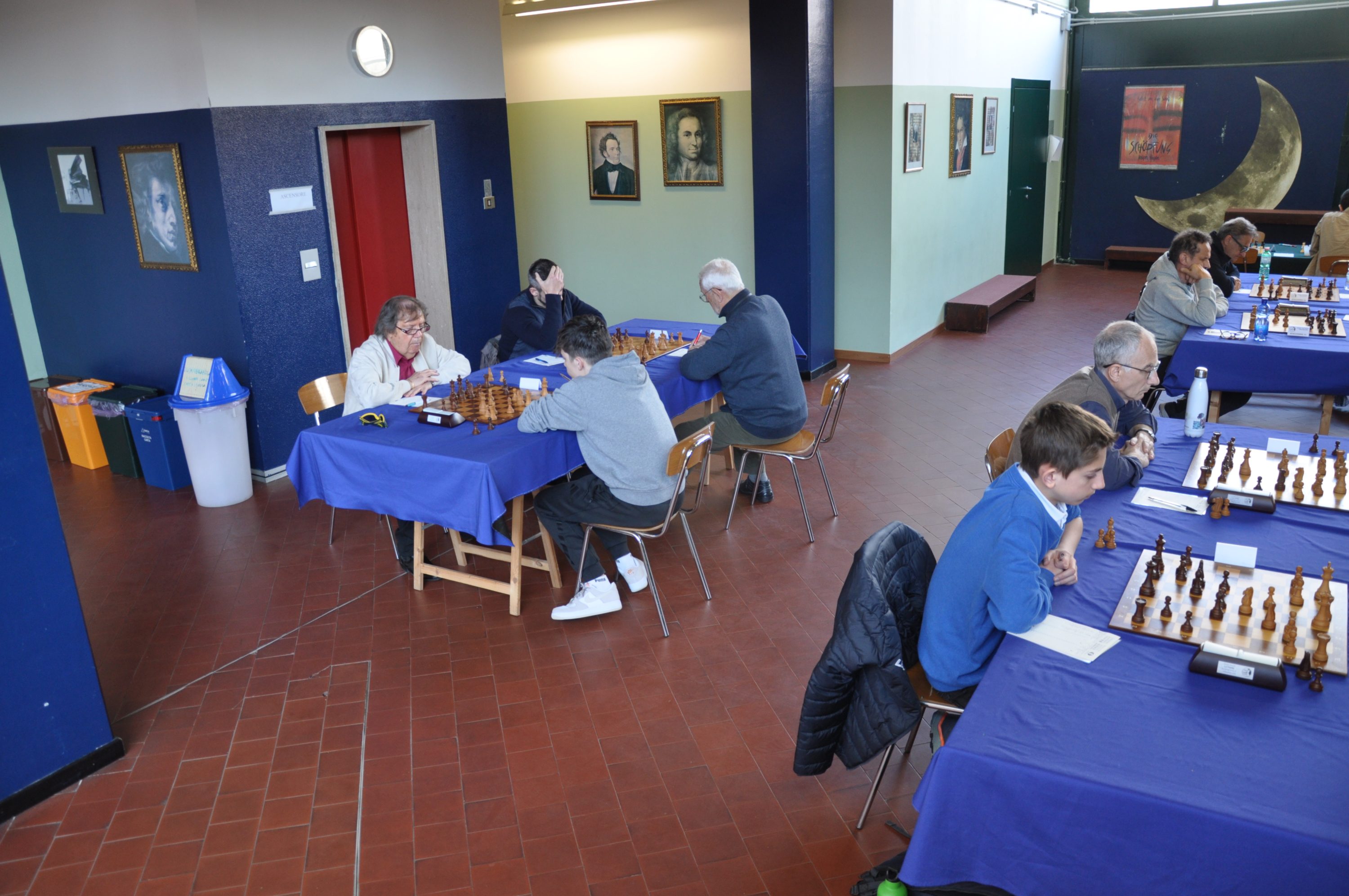 A wide view of a chess tournament taking place indoors. Several participants are seated at tables covered with blue tablecloths, focused on their chess games. Paintings adorn the walls in the background, and a clock is visible above.