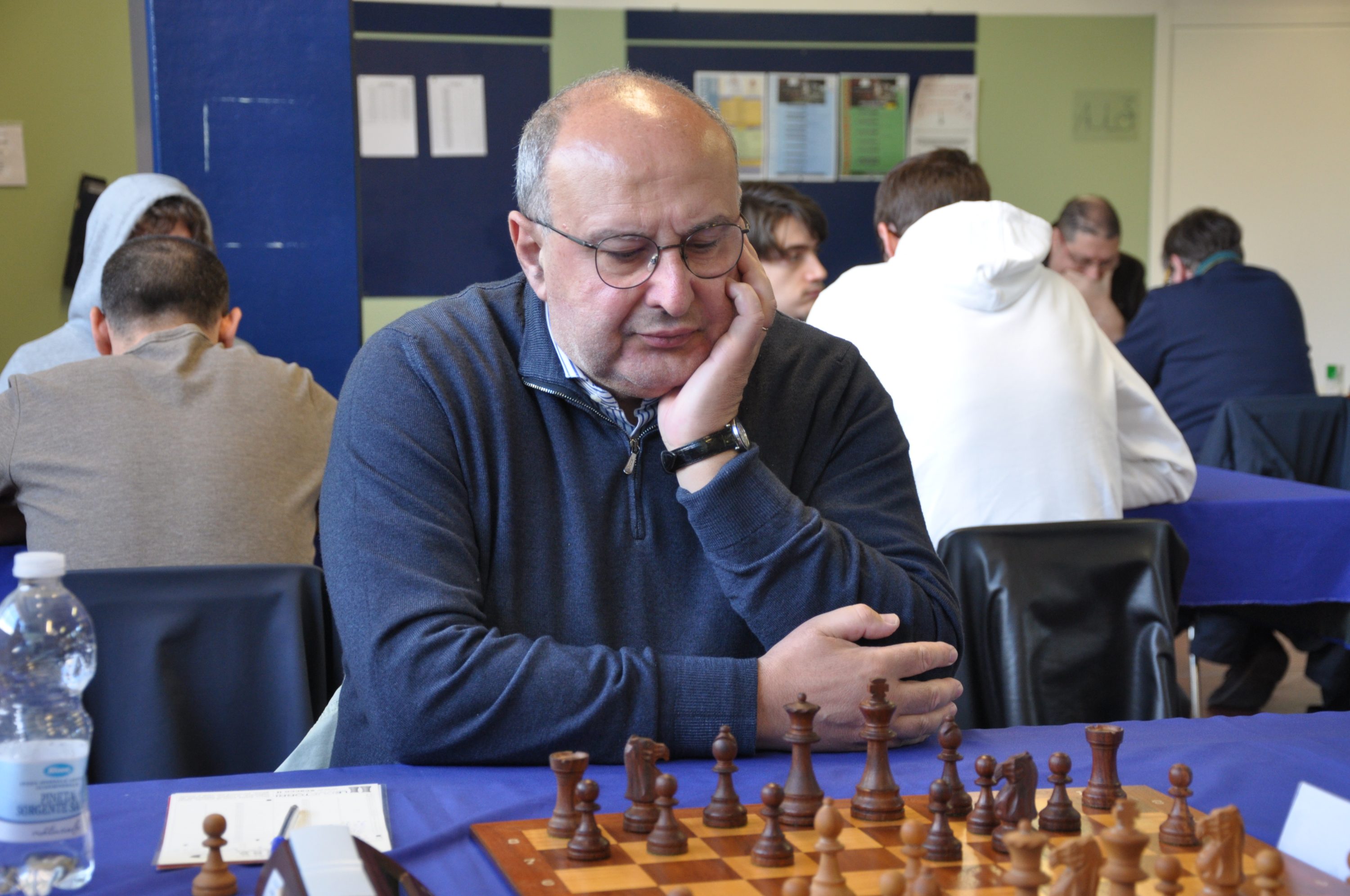 A contemplative man with glasses sits at a chess table, focusing intently on the game. Chess pieces are set up in front of him, while other players can be seen in the background.